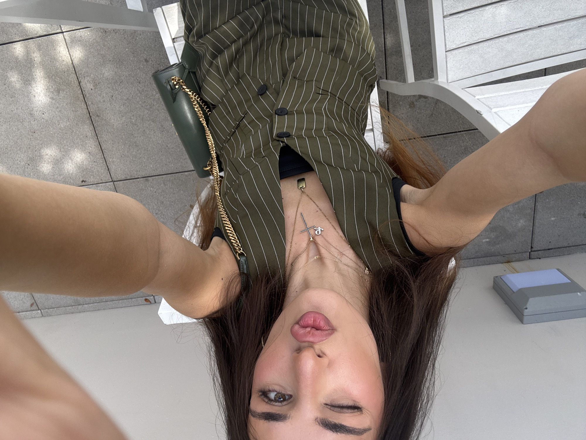 In Hallandale Beach, Florida, a young woman is captured in a selfie outdoors, seated on a white chair. She is wearing a green pinstripe vest over a black top and accessorized with multiple gold necklaces, one featuring a cross pendant. A green crossbody bag with a gold chain strap is visible on her right side. The woman is winking and puckering her lips, looking towards the camera. The background shows a light-colored ceiling, a grey outdoor light fixture, and grey paving stones.