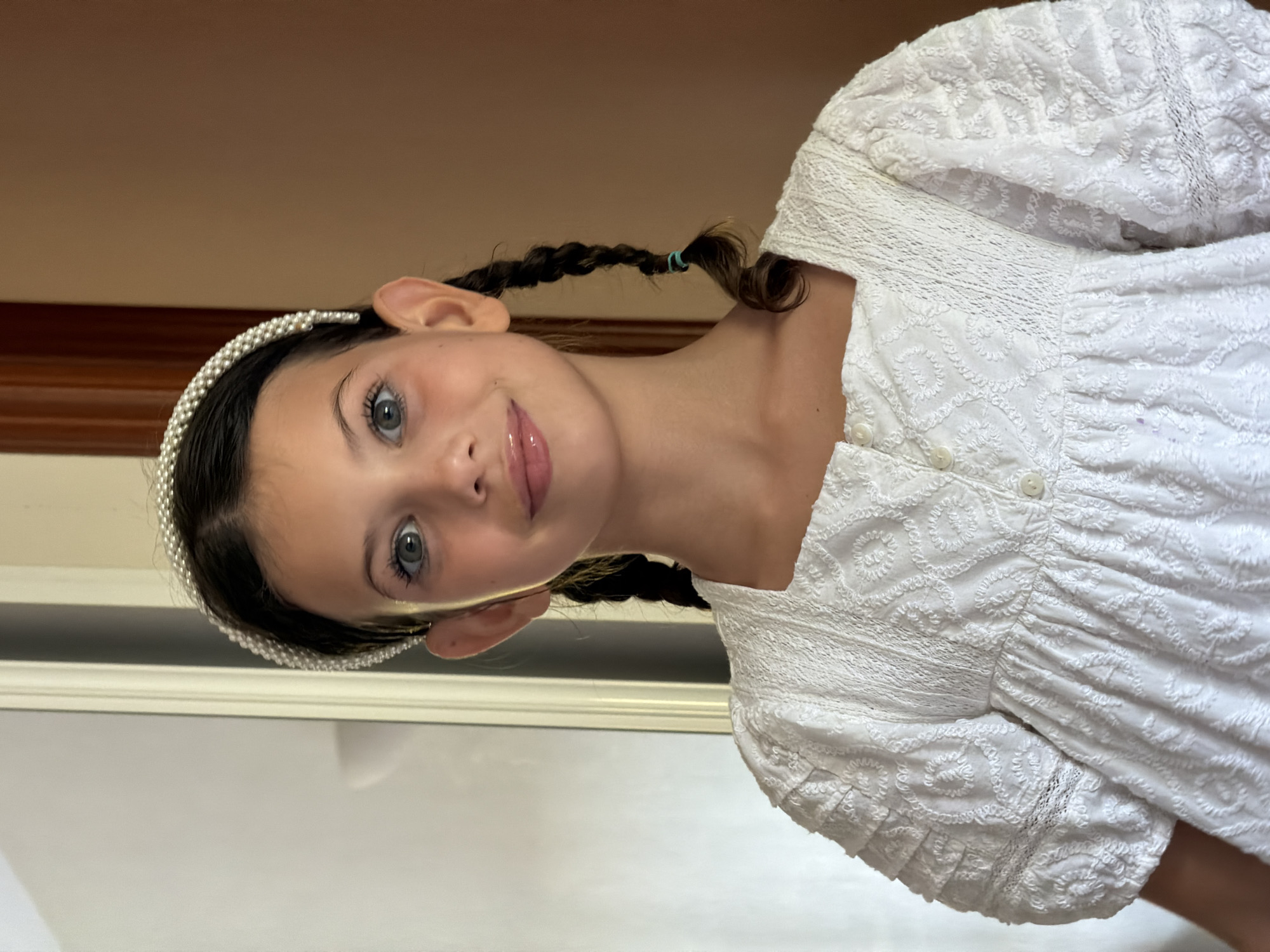 A young girl with dark hair styled in two braids, wearing a white dress and a pearl headband, smiles at the camera. She is positioned in the center of the frame, with a blurred background that includes a doorway and a wall. The lighting is soft and natural, suggesting an indoor setting.