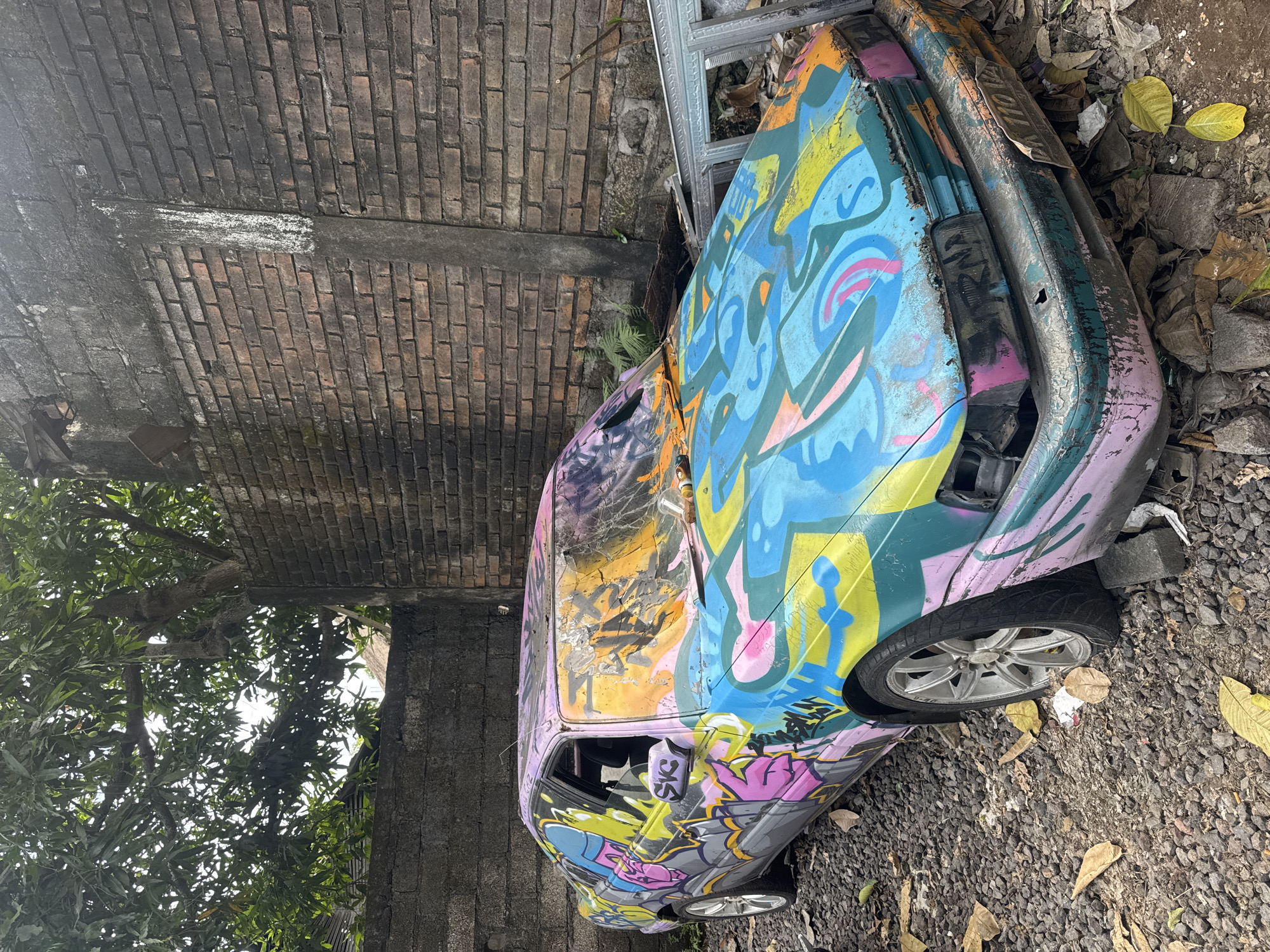 An abandoned, graffiti-covered sedan sits in a lot filled with gravel and debris. The car, a muted teal with large swaths of pink and purple paint, is heavily vandalized with bright, abstract graffiti in shades of blue, yellow, orange, and pink. The windshield is shattered and covered in more paint. The car appears to be an older model, possibly a Mazda 323, with its front bumper damaged and parts of its interior exposed. The background consists of a weathered brick wall and overgrown vegetation, suggesting an urban or semi-urban environment. A license plate, partially obscured, is visible on the front of the car, reading "DK 1102 PQ," suggesting it is registered in Bali, Indonesia. The scene depicts a neglected and deteriorated vehicle, transformed into a canvas for street art in Kuta Selatan, Indonesia.