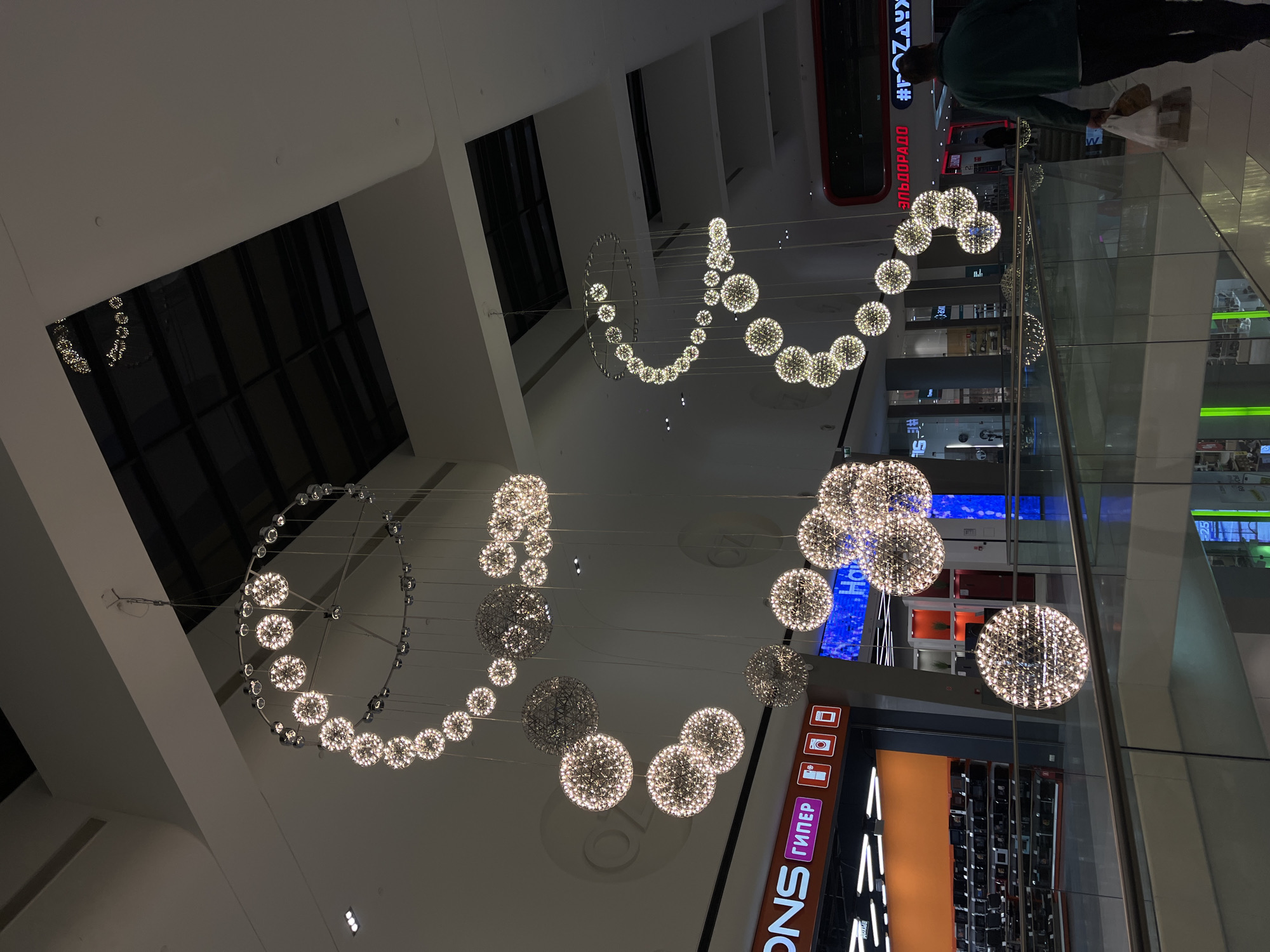 The image displays the interior of a large, modern building, likely a shopping mall, characterized by its high ceilings and elaborate decorative lighting. Numerous spherical chandeliers, crafted from many small, glittering lights, hang suspended, forming dynamic patterns that include elongated waves and complete circles. These fixtures provide the primary illumination for the space.
One person is visible in the upper right quadrant of the image, seen from behind. They are dressed in a dark green top and dark pants, and are carrying a light-colored bag, suggesting they are a shopper or visitor moving through the facility. The perspective implies the person is on an upper level, looking down or across the expansive interior.
Glass railings are prominent features, indicating a multi-story structure with various walkways or balconies overlooking the main area. In the background, multiple shop fronts and displays are discernible, reinforcing the commercial nature of the setting. The lighting is entirely artificial, creating a bright yet focused ambiance typical of an indoor public space, irrespective of the time of day outside.
Several Russian brand names are visible on store signage. These include "М.ВИДЕО" (M.VIDEO) and "#Эльдорадо" (Eldorado), both identifiable as major electronics retailers. Another sign clearly shows the word "гипер" (giper), which is likely an abbreviation for "гипермаркет" (hypermarket). The scene captures a typical moment within a modern commercial hub in Tliustenkhabl', Russia.
