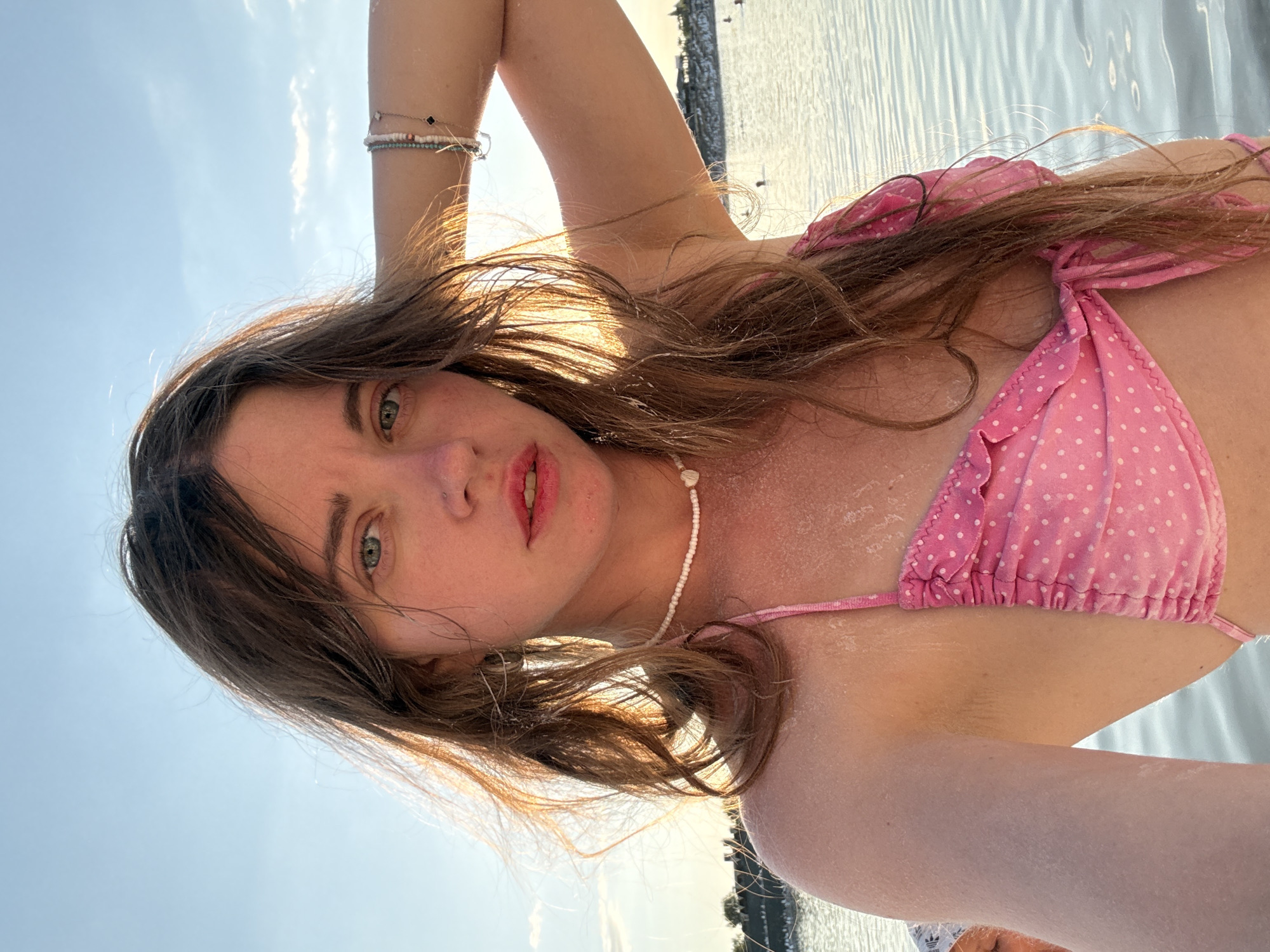 A selfie taken by a young woman with long, wavy brown hair and blue eyes, wearing a pink polka dot bikini. She is standing in shallow water under a clear blue sky with the sun setting. The water is calm and reflects the sky. In the background, there is a distant shoreline with some buildings. The woman has her left arm raised, touching her hair, and is looking directly at the camera. She is wearing a white beaded necklace with a heart pendant. The overall scene is serene and picturesque, suggestive of a warm, summer day by the water in Sol-Iletsk, Russia.