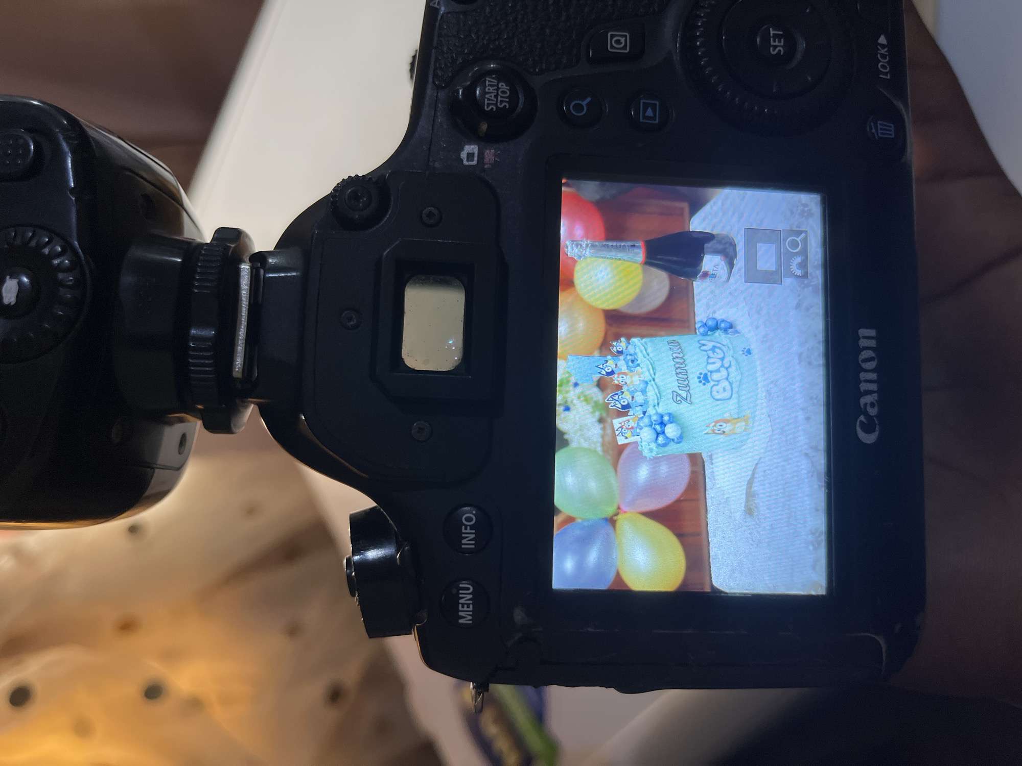 A close-up, angled shot captures the rear screen of a black Canon DSLR camera. The camera's screen displays an image of a light blue cake decorated with cartoon characters and the name "Zunmar" in blue lettering. The cake is adorned with blue balloons and a small bottle of sparkling wine. In the background of the screen image, several colorful balloons are visible. The camera itself is partially obscured by its flash unit, which is extended upwards. The ambient lighting in the room is warm and soft, casting an orange glow on a sheer fabric in the upper left corner. The background suggests an indoor setting, possibly a home or event space. The camera has several buttons and controls visible, including "MENU," "INFO," "START/STOP," and "SET." The "Canon" logo is visible on the bottom edge of the camera's screen. The image does not contain any information indicating a location in Bukuru, Nigeria.