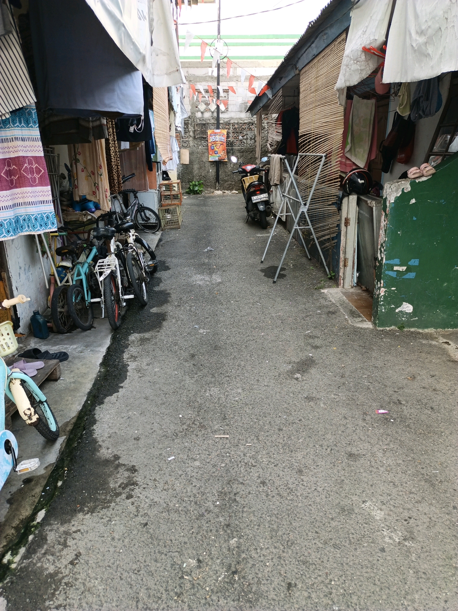 The image depicts a narrow, paved alleyway in what appears to be a densely populated residential area, likely a kampung or urban slum, in South Jakarta, Indonesia. The alley is flanked by simple, informally constructed buildings made of wood, bamboo, and corrugated metal, with tarpaulins and fabrics draped over their fronts, suggesting residential dwellings or small shops. Laundry is hung out to dry from the structures.
Several bicycles are parked along the left side of the alley, leaning against the buildings and a makeshift rack. One light blue bicycle is partially visible in the foreground. A red and yellow scooter is parked on the right side of the alley, in front of a building with bamboo matting. A metal drying rack stands in the middle of the alley.
A poster, possibly an advertisement, is affixed to a wall on the right side of the alley. Red and white Indonesian flags are strung across the alleyway above, indicating a potential national celebration or event. The ground is asphalt with patches of wetness and debris, indicative of an outdoor environment. The overall impression is one of a lived-in, functional space within a bustling urban setting. There are no people visible in the image.