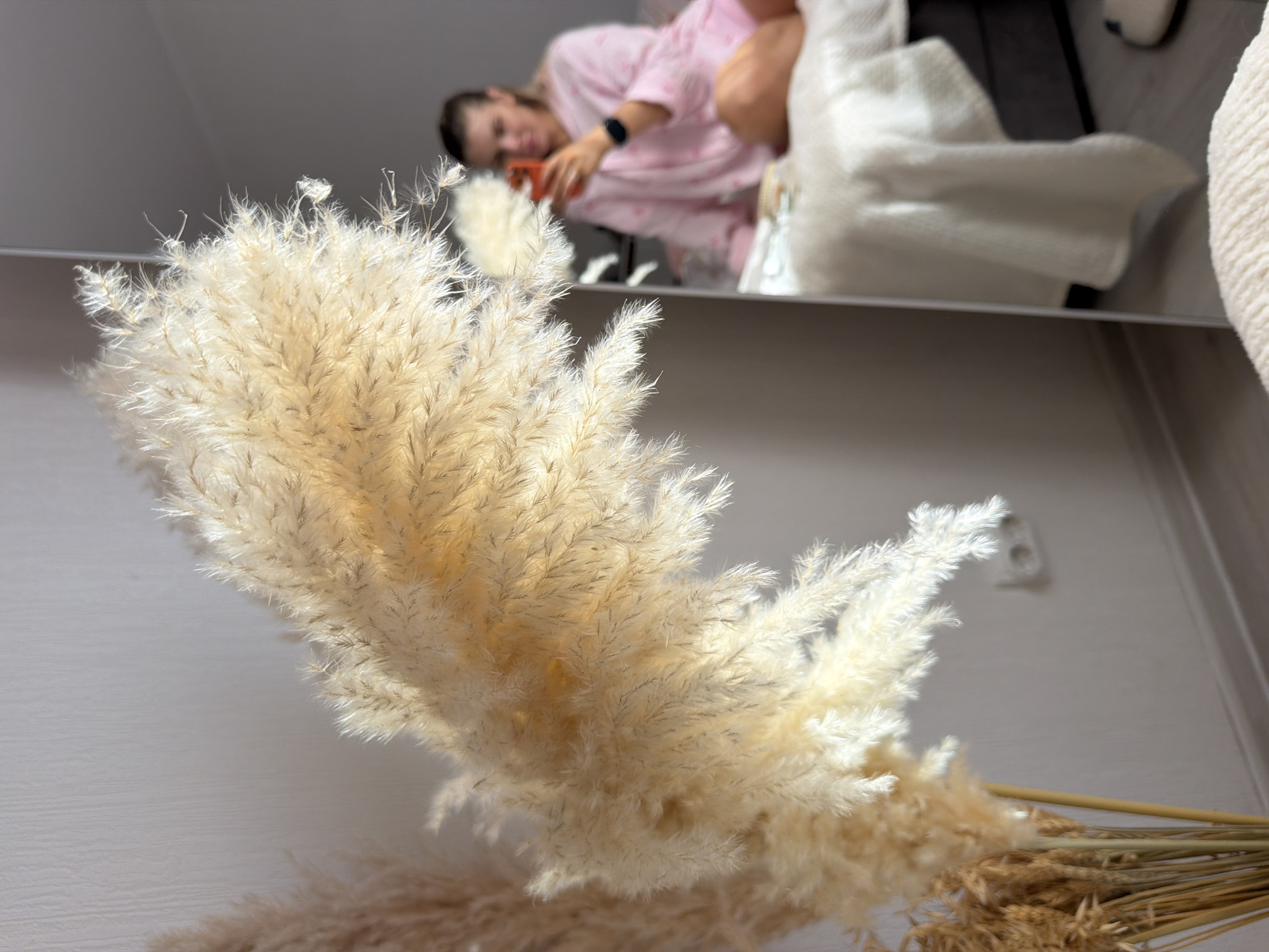 This image shows a close-up of dried pampas grass plumes in the foreground, with a reflection in a mirror to the right. In the reflection, a person is visible, seated on a bed and holding a smartphone. The person is wearing a pink long-sleeved pajama set with a pattern. The background environment appears to be a bedroom, with a white blanket on the bed and a light-colored wall. There is no discernible information in the image to indicate the location is Moscow, Russia.