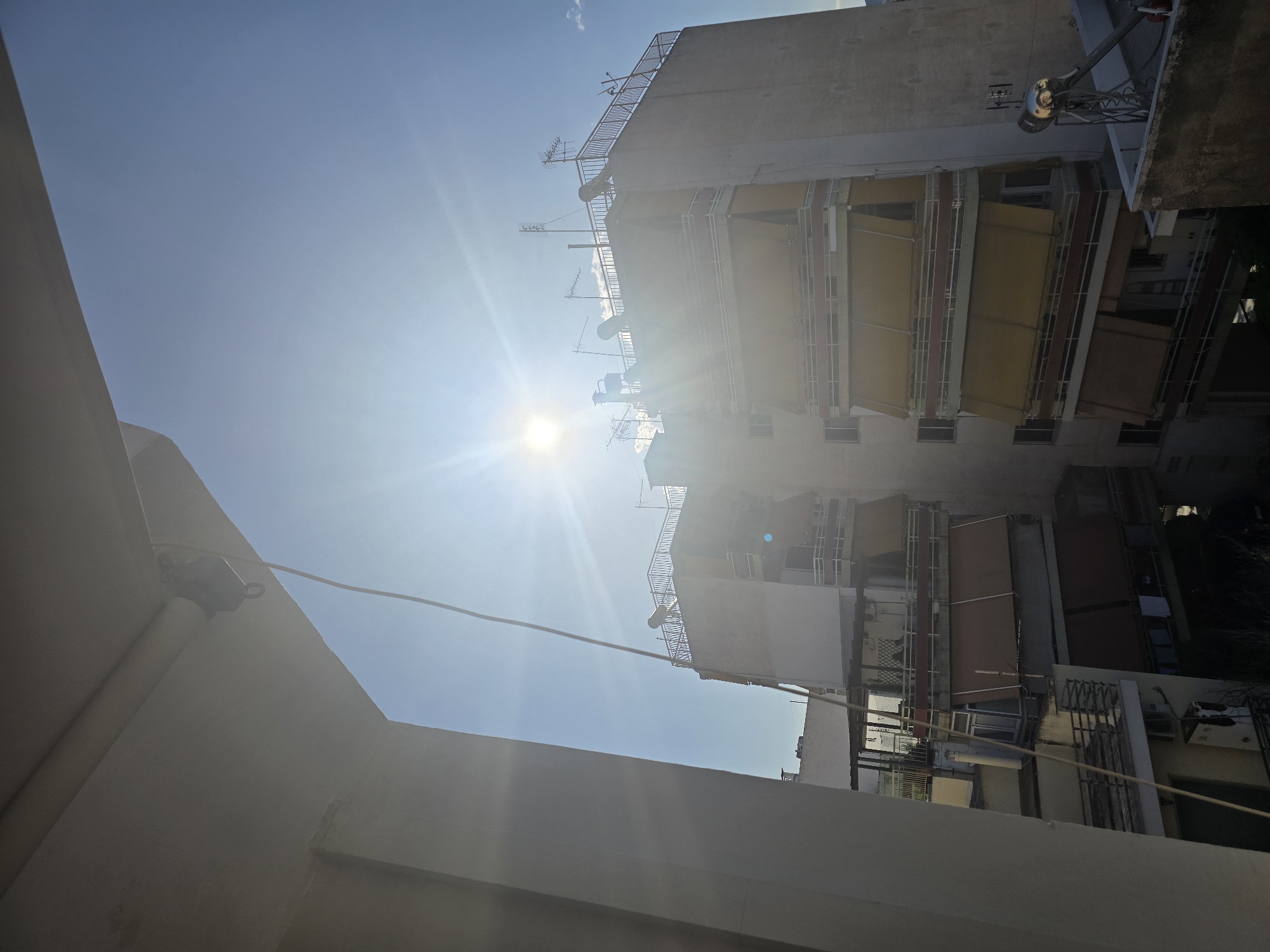 The image shows a view of apartment buildings in Agii Anargiri, Greece, on a sunny day. The sun is bright and positioned high in the sky, creating lens flare and strong highlights. A prominent white building facade, possibly part of a balcony or overhang, is in the foreground on the left, partially obscuring the view of the sky and the buildings beyond. A thin wire hangs vertically from the overhang into the frame.
The main subject is a multi-story apartment building with multiple balconies. Many of the balconies are equipped with retractable awnings, most of which are extended and are a yellowish-tan color. Some awnings are retracted or not visible. The building has white railings on its balconies and appears to be constructed of concrete. The rooftop of the building is visible and is equipped with several antennas, including a large satellite dish and smaller aerials. There are also what appear to be solar water heaters on the rooftop. Another, slightly shorter building is visible behind and to the left of the main apartment building. The sky is clear and blue, with a few wispy clouds visible. The overall scene depicts an urban residential environment. No people are visible in the image.