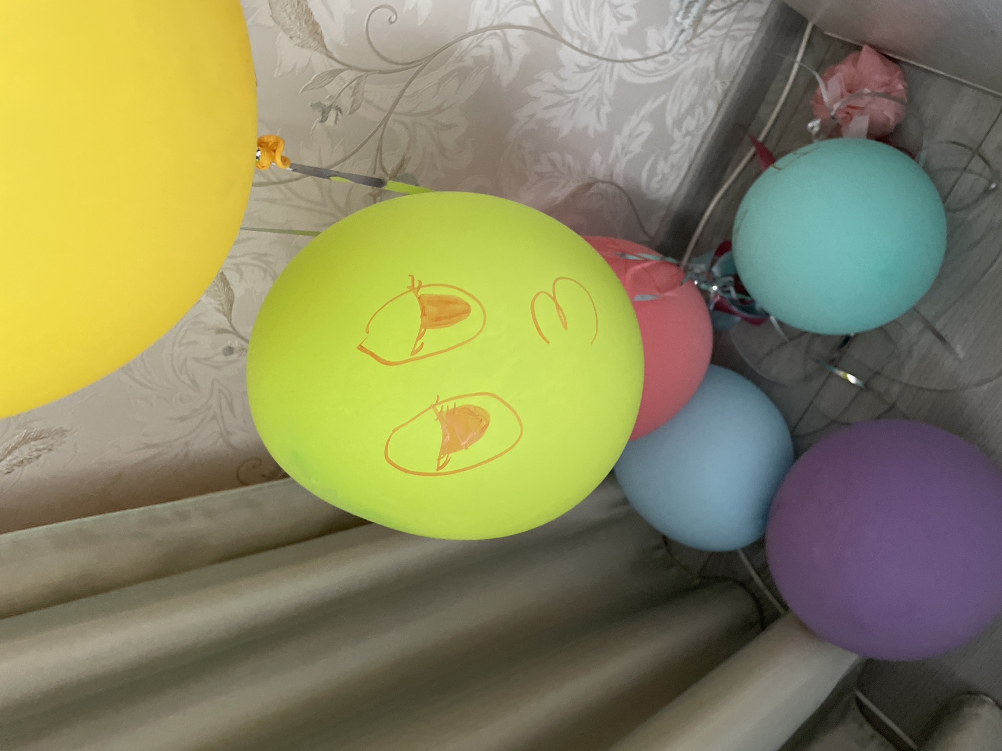 The image shows a collection of balloons of various colors, including yellow, light green with a drawn face, pink, light blue, and purple. They are likely party decorations. A segment of a curtain is visible in the lower portion of the image. The backdrop appears to be a patterned wallpaper, possibly floral. Based on reverse image searches, the image may originate from Moskovskiy, Russia.