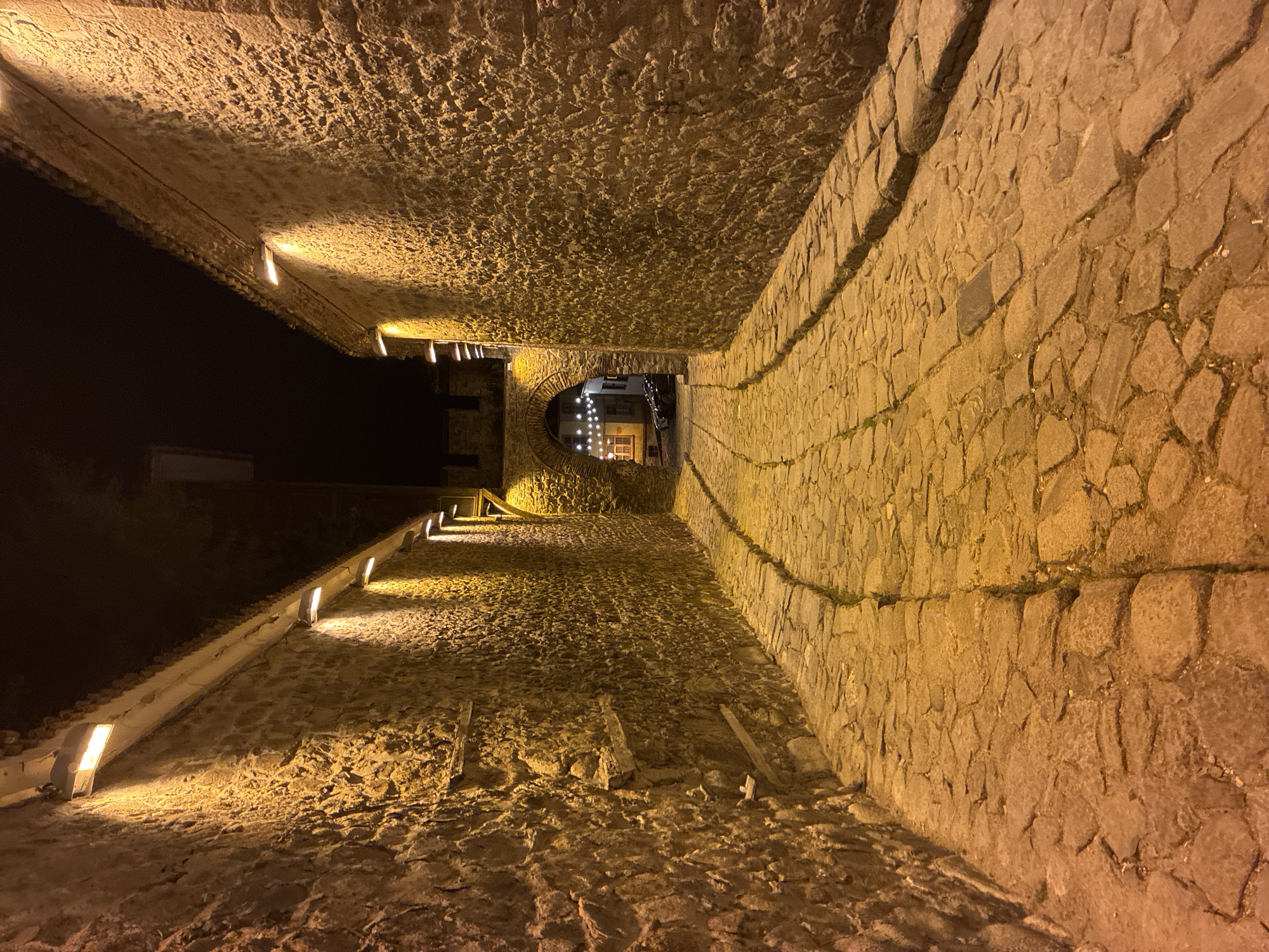 The image depicts an upward-sloping cobblestone pedestrian pathway in Plovdiv, Bulgaria, at night. Stone walls flank both sides of the pathway, with built-in lights illuminating the route. An archway is visible in the distance at the end of the path, beyond which a cluster of lights hangs above the street. The sky is black.