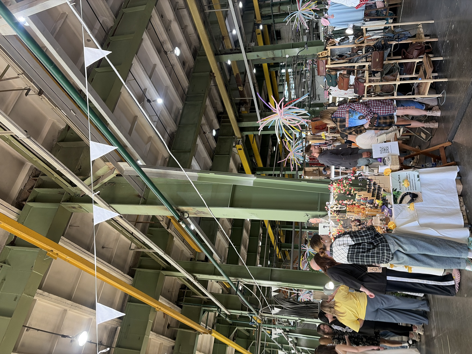 This image captures an indoor market scene within a large, repurposed industrial hall at Sevkabel Port in Saint Petersburg, Russia. The building features an expansive ceiling supported by heavy olive-green painted steel columns and beams. Overhead, prominent yellow crane rails are visible, spanning the length of the hall, alongside white concrete ceiling panels, industrial lighting fixtures with exposed bulbs, and various utility conduits. A string of white triangular pennant flags is stretched across the upper portion of the scene, and several colorful, starburst-like decorative elements hang down.
In the midground and foreground, numerous market stalls are arranged, displaying various goods. The stalls consist of tables covered with white tablecloths. On the left, a group of people, predominantly young adults in casual attire, are browsing. One woman wears a yellow t-shirt and grey trousers, while another is in a black top with white-striped trousers. Towards the center-right, a woman wearing a plaid shirt and light-wash jeans stands in front of a stall laden with jewelry, including necklaces, bracelets, and small decorative items. A sign on this table is partially visible, indicating "Ароналава," likely a brand name. To the right of this stall, a multi-tiered wooden display stand showcases an assortment of leather handbags and accessories. A man in a dark grey shirt stands behind this display, while another woman in a plaid jacket holds a book or tablet near the handbag display. The floor is a dark, possibly concrete, surface. The scene depicts a lively market environment with individuals observing and selecting items.