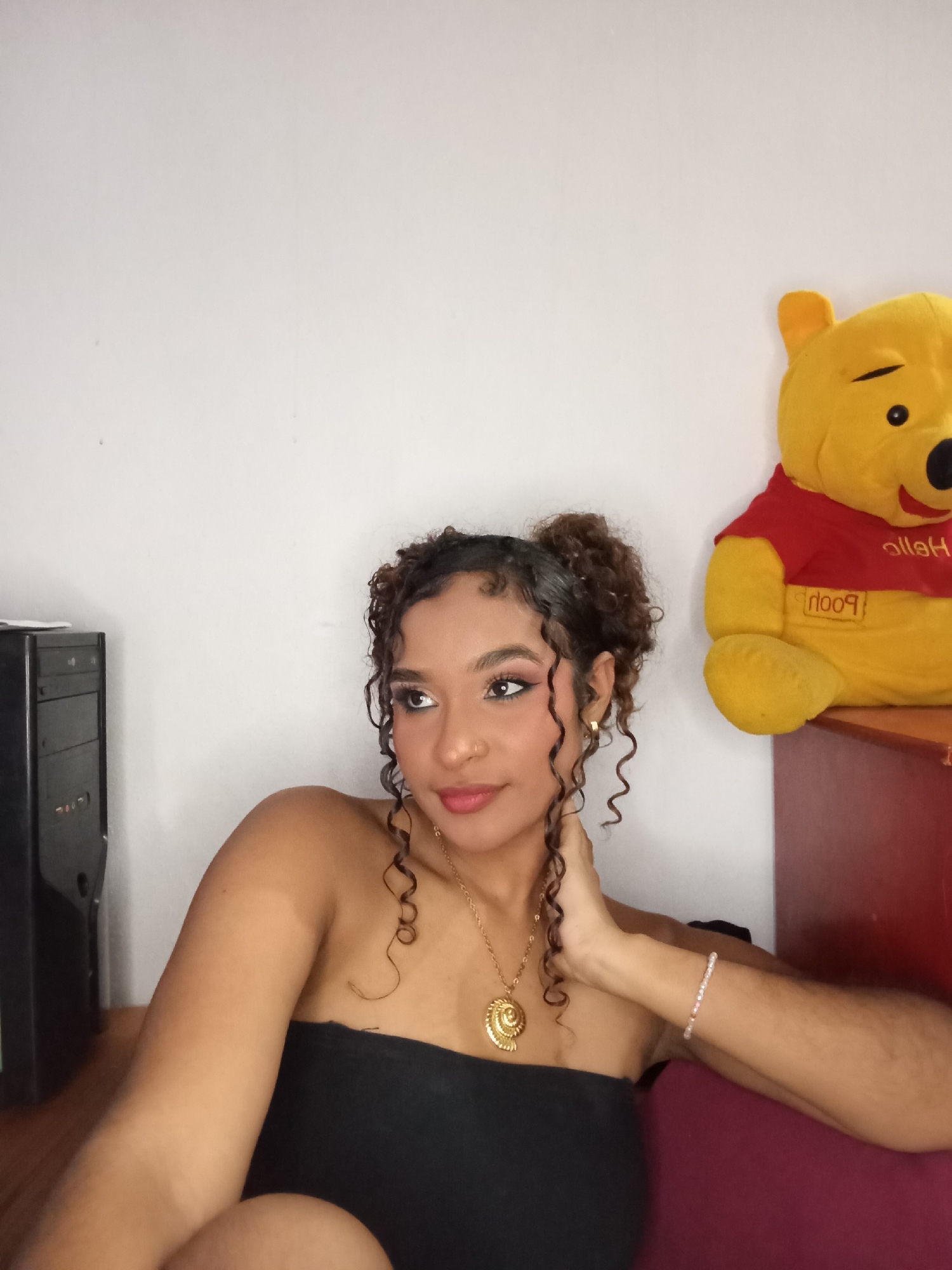 The image displays an indoor scene in Juan Griego, Venezuela, featuring a young adult female seated in front of a light-colored wall. The female has light brown skin, dark curly hair styled in two top buns with loose curls framing her face. She wears winged eyeliner, light eyeshadow, mascara, and pink lipstick, a nose stud, and small gold hoop earrings. Her attire consists of a black strapless top, a gold chain necklace with a shell-shaped pendant, and a light-colored beaded bracelet on her right wrist. Her right hand is positioned at her neck/jawline as she gazes towards the upper left portion of the frame with a subtle smile. To the left and slightly behind her, a black computer tower is visible. To her right, a yellow Winnie the Pooh stuffed animal, wearing a red shirt with "Pooh" inscribed, sits on a dark red/maroon surface, likely a shelf or cabinet. The female herself is seated on a piece of furniture upholstered in a similar dark red/maroon fabric.