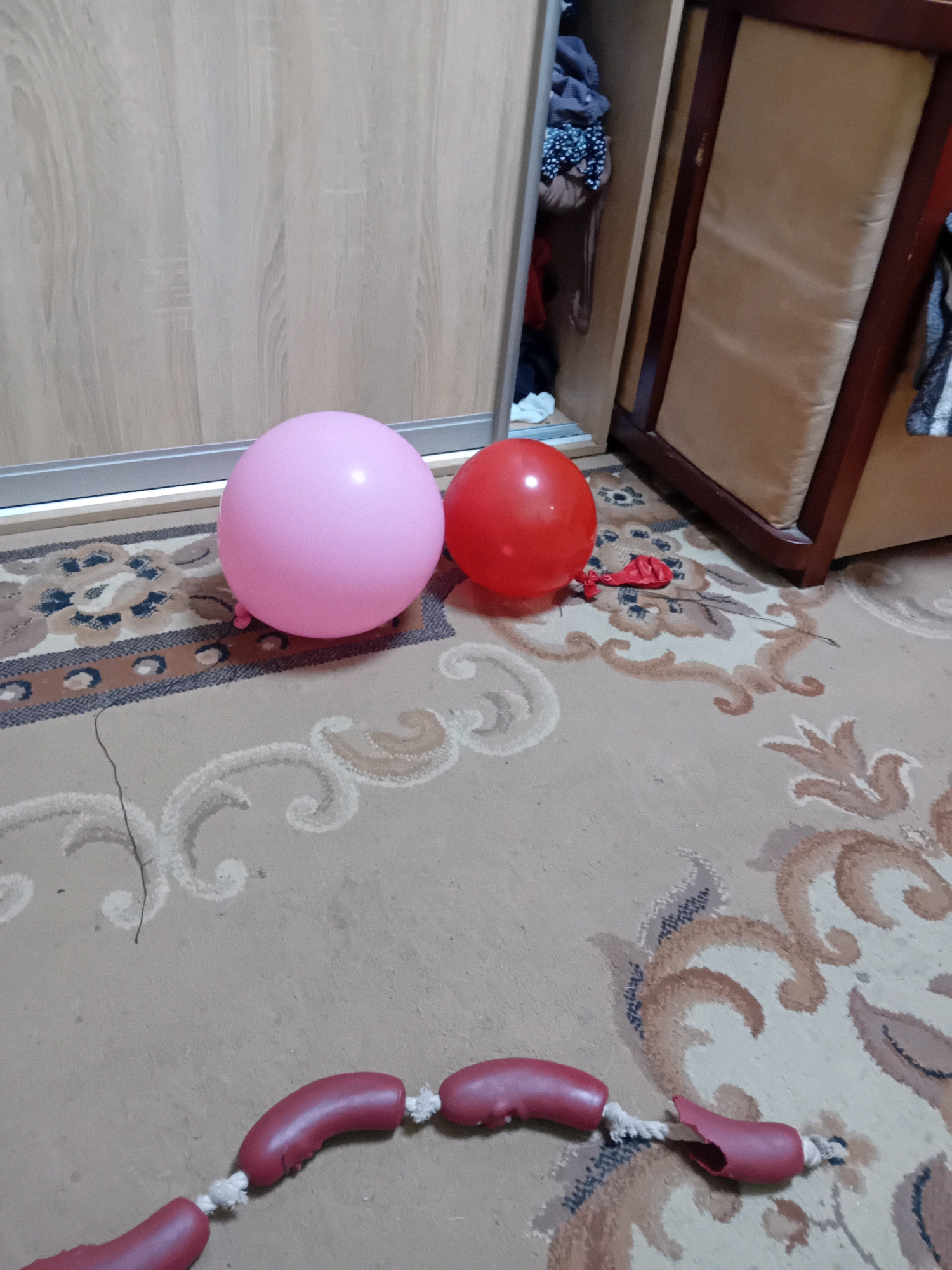 A pink balloon and a red balloon lie on a patterned carpet in front of a wooden closet and a wooden piece of furniture with a beige upholstered seat. A string of pink, sausage-shaped objects is also on the carpet in the foreground. The scene is indoors.