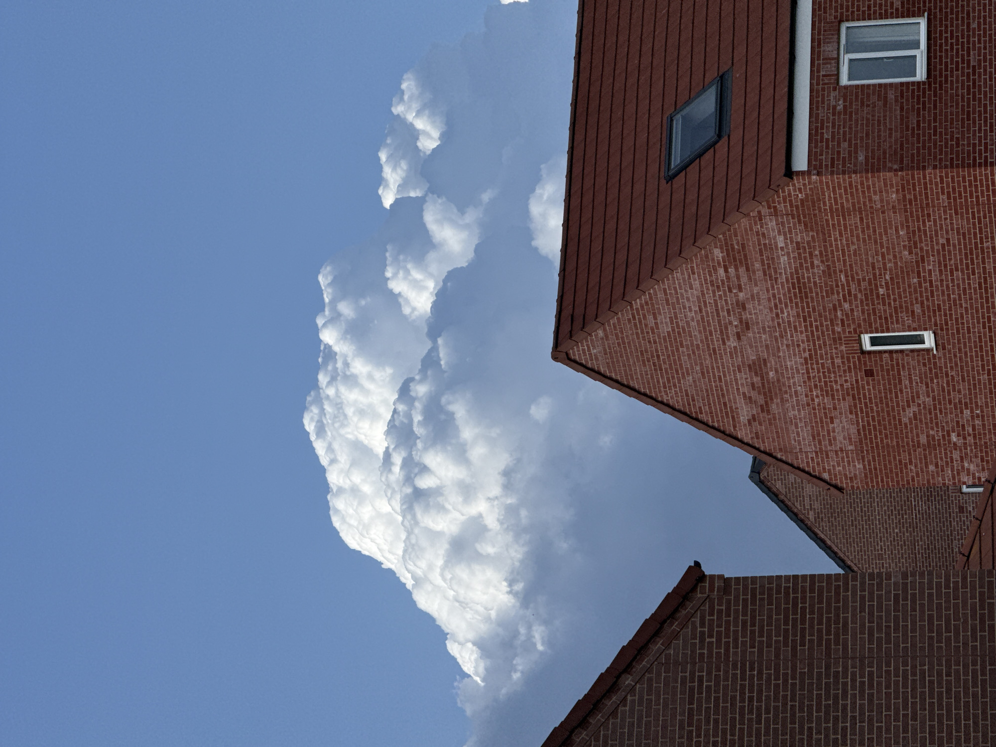 The image shows a low-angle view of red brick houses in Great Yarmouth, United Kingdom, against a sky with a large, towering cumulonimbus cloud formation. The houses have pitched roofs with dark red shingles. One house displays two rectangular windows, one larger than the other, on the visible wall. The sky transitions from clear blue to areas obscured by the massive cloud.