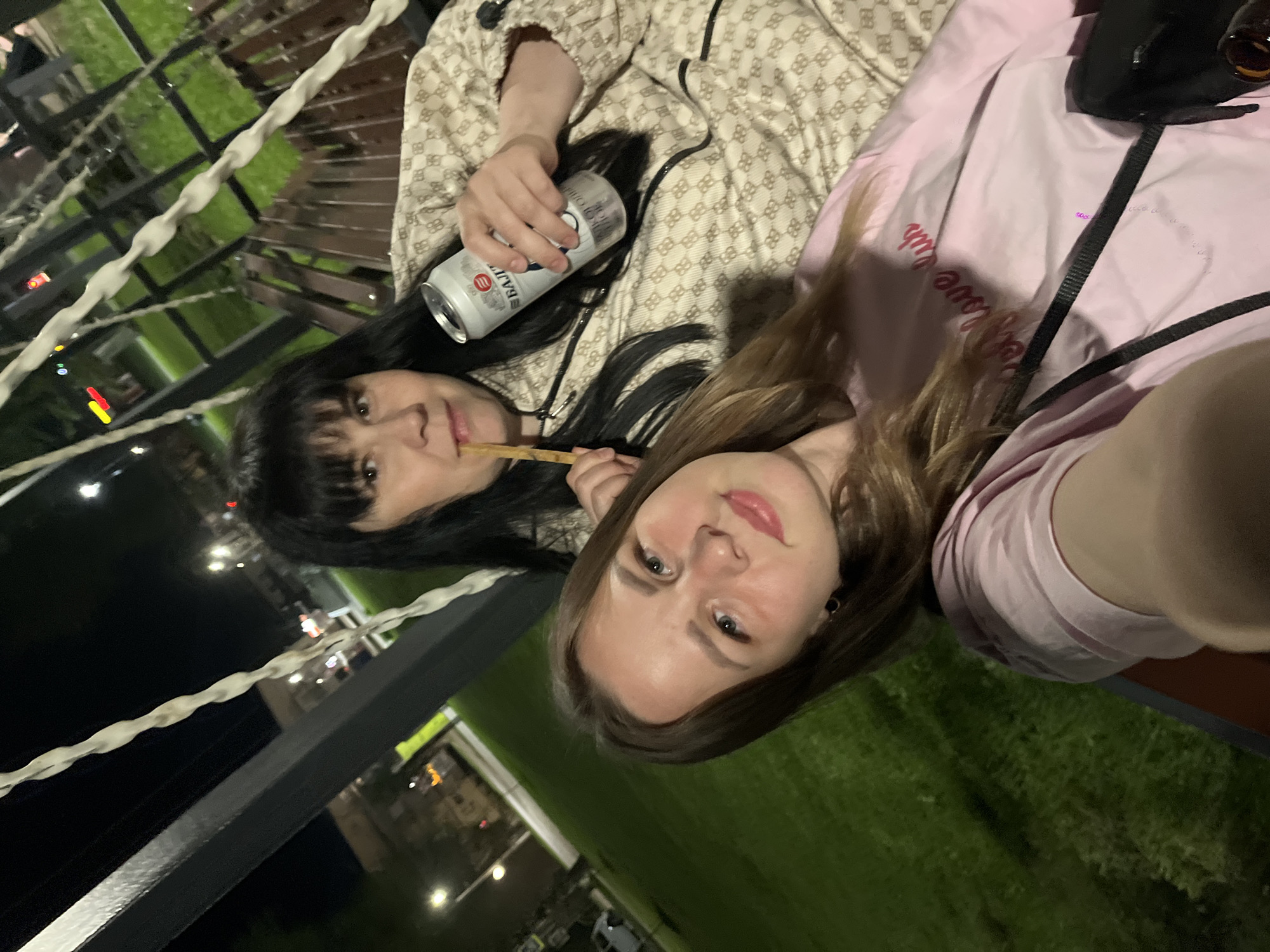 Two women are posing for a selfie outdoors, likely in Zav'yalovo, Russia. The older woman has dark hair with bangs, is wearing a patterned jacket, and holding a can of Baltika beer. She appears to be holding a wooden stick in her mouth. The younger woman has long, light brown hair and is wearing a pink top with text on it. The background suggests an outdoor setting with a metal railing, greenery, and city lights in the distance.