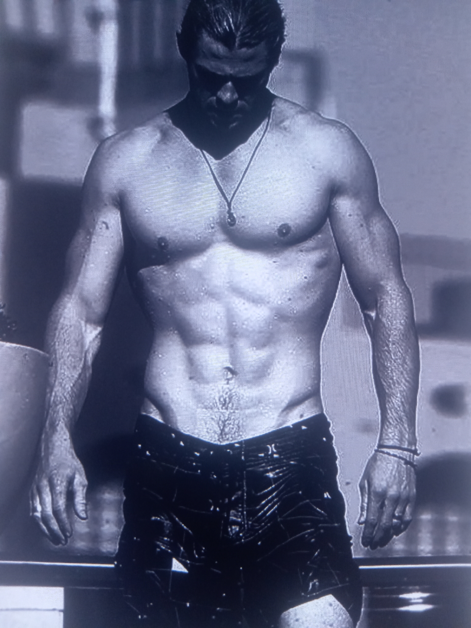 A black and white, medium shot photograph shows a muscular man standing in water, possibly a swimming pool. The man is shirtless, wet, and wearing dark shorts with a textured pattern. His abs are clearly defined, and he has water droplets on his skin. He is wearing a necklace with a pendant. His arms are at his sides, and his hands are visible. The background is blurred and appears to be an outdoor setting, possibly with some structures and sunlight. The man's face is shadowed, but he appears to be looking downwards.