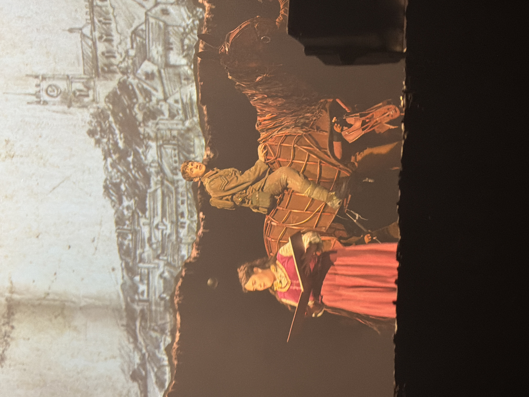 The image depicts a scene from a theatrical production, possibly projected onto a surface. A man in military attire is lying on a structural piece that resembles a large, woven basket or part of a wicker animal. A woman in period clothing, featuring a pink dress with a golden-trimmed bodice, holds an object (possibly a prop sword or a staff). In the background, there is a depiction of an older architectural design or map that is reminiscent of historic London. It is not possible to definitively identify a specific location in London from the image provided.