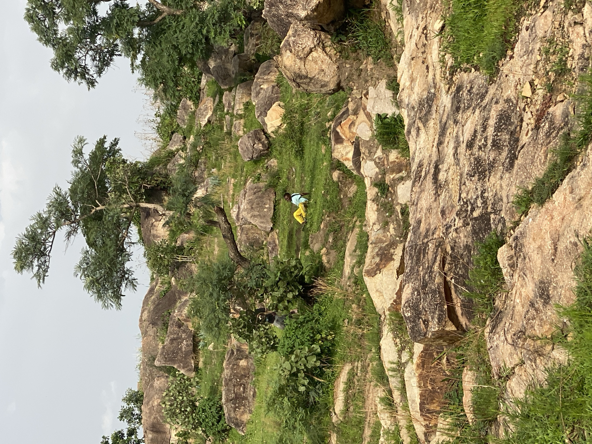 In the rocky terrain of Du, Nigeria, two individuals are observed. One person, wearing yellow pants and a light-colored top, is seated on a grassy slope amidst large, weathered rocks. Another person, dressed in dark clothing, is partially obscured by dense green foliage to the left of the seated individual. The environment is characterized by rugged, boulder-strewn hills dotted with sparse trees and green vegetation under a hazy sky.