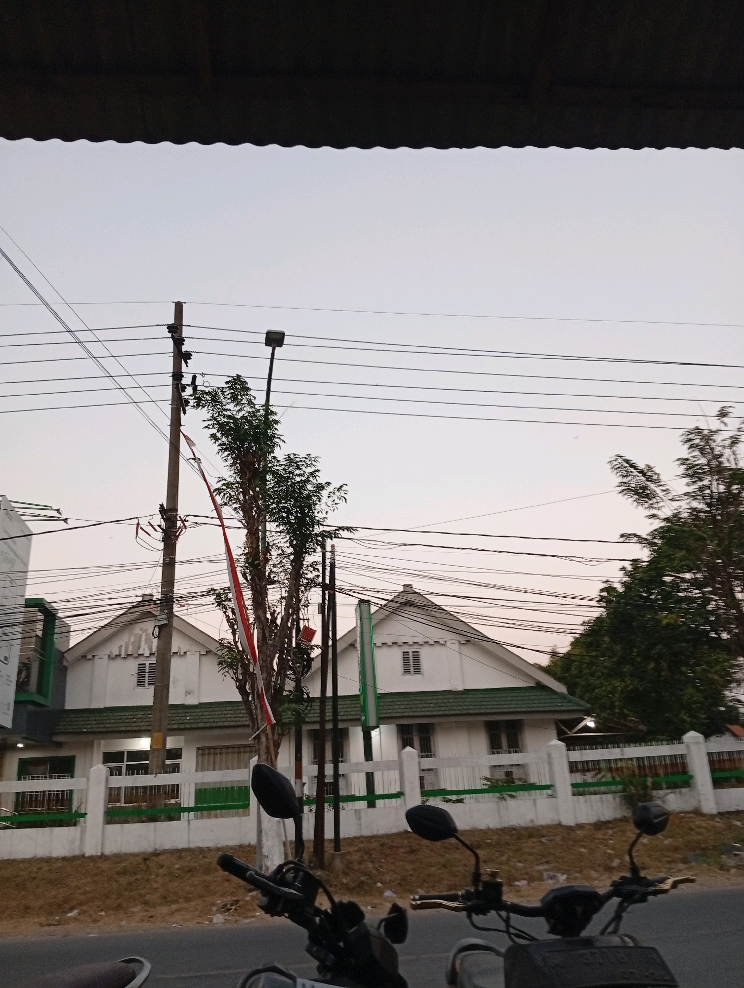 The image shows a street scene in Banyu Ajuh, Indonesia. In the foreground, the handlebars and part of the front of a black motorcycle are visible. The motorcycle is parked on a paved road, with a grassy embankment to its right. Beyond the embankment, a white fence with green accents runs along the front of a building. The building appears to be a single-story structure with a green roof and several windows. A tall utility pole with numerous electrical wires is situated to the left of the building. A tree with a red and white Indonesian flag draped over it stands near the utility pole. The sky is overcast and a dark, wavy overhang is visible at the top of the image. The overall scene suggests an outdoor, urban environment.