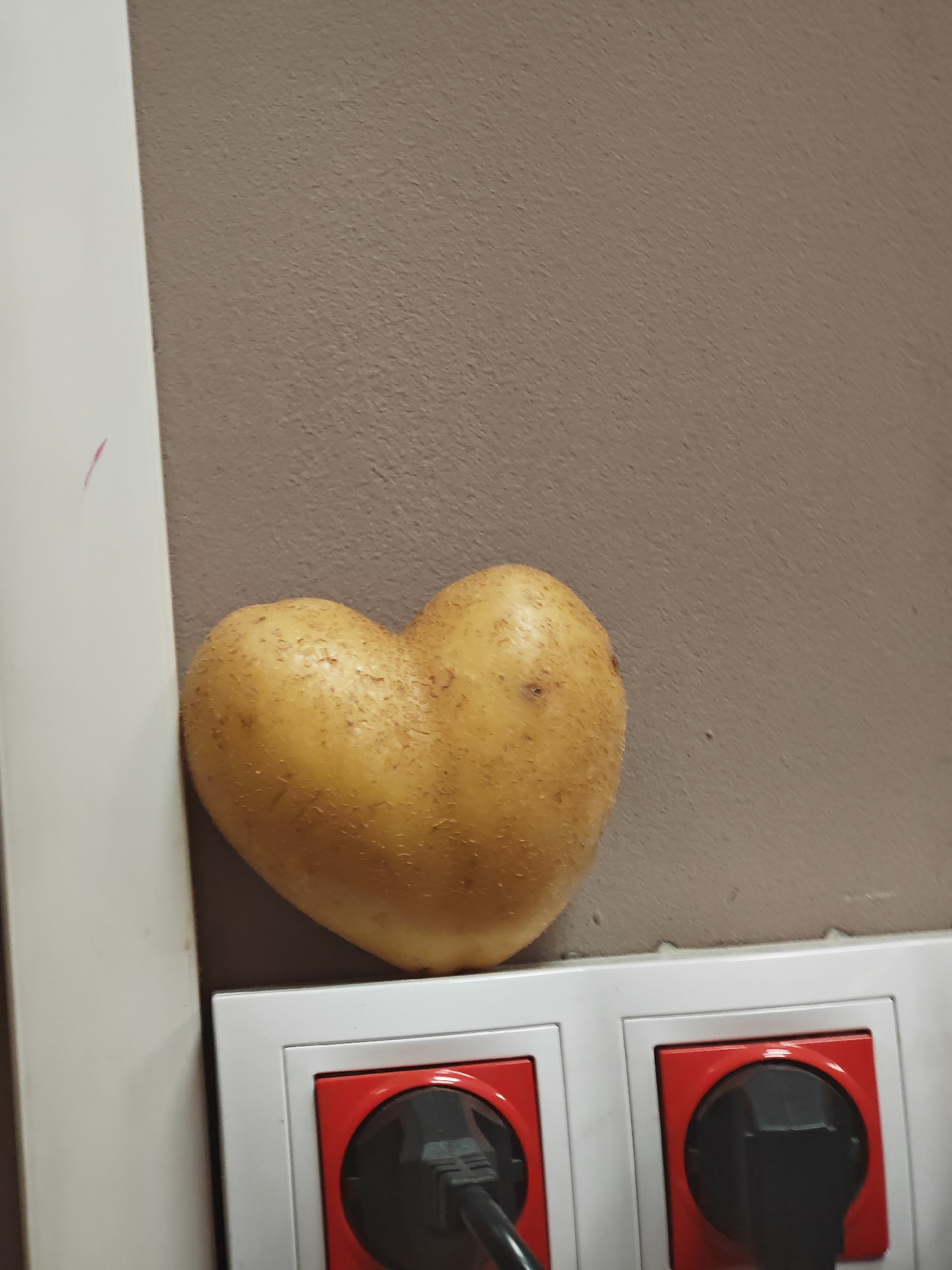 In an indoor setting within Shumen, Bulgaria, a naturally heart-shaped potato with light brown, textured skin is positioned atop a white multi-socket electrical outlet strip. The potato leans against a textured, matte beige wall. The outlet strip contains at least two visible sockets, each featuring a prominent red internal housing. A black electrical plug is inserted into the left socket, while the right socket remains empty. A vertical white architectural element, likely a door frame or trim, is partially visible along the image's left edge, displaying a faint, vertically oriented pink mark on its surface. No individuals or active interactions are depicted.