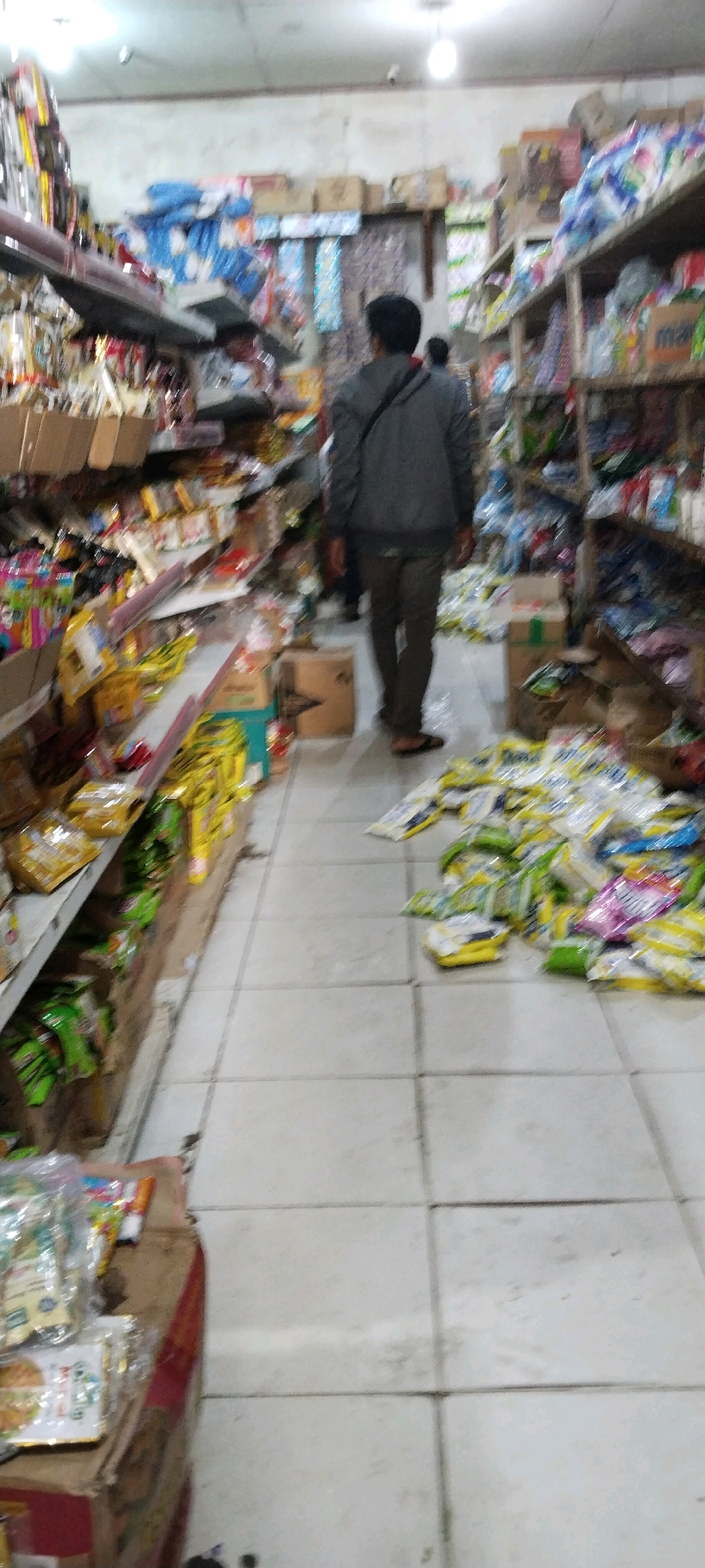 A poorly lit, low-resolution image shows the interior of what appears to be a small grocery store in Bandar Jaya, Indonesia. The aisle is lined with shelves stocked with various goods, and products are also scattered on the floor, particularly on the right side of the image, suggesting disarray or a recent incident. A man in a gray jacket and dark pants is walking away from the camera, his back to the viewer, down the aisle. Another person, partially visible, is walking ahead of him. The flooring is tiled, and the walls are painted a pale color. The overall impression is one of a cluttered and somewhat unorganized retail space.