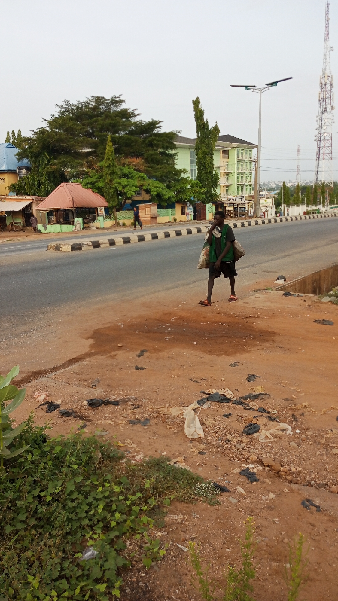 The image shows a man walking along a road in Adankolo, Nigeria. He is carrying two large bags over his shoulders. He is wearing a green vest, dark shorts, and sandals. The road is lined with black and white traffic barriers. Across the road are buildings, including a green-painted multistory building and a small structure with a red roof. A communication tower is visible in the distance. The shoulder of the road is dirt and littered with trash. Vegetation grows at the bottom of the frame.