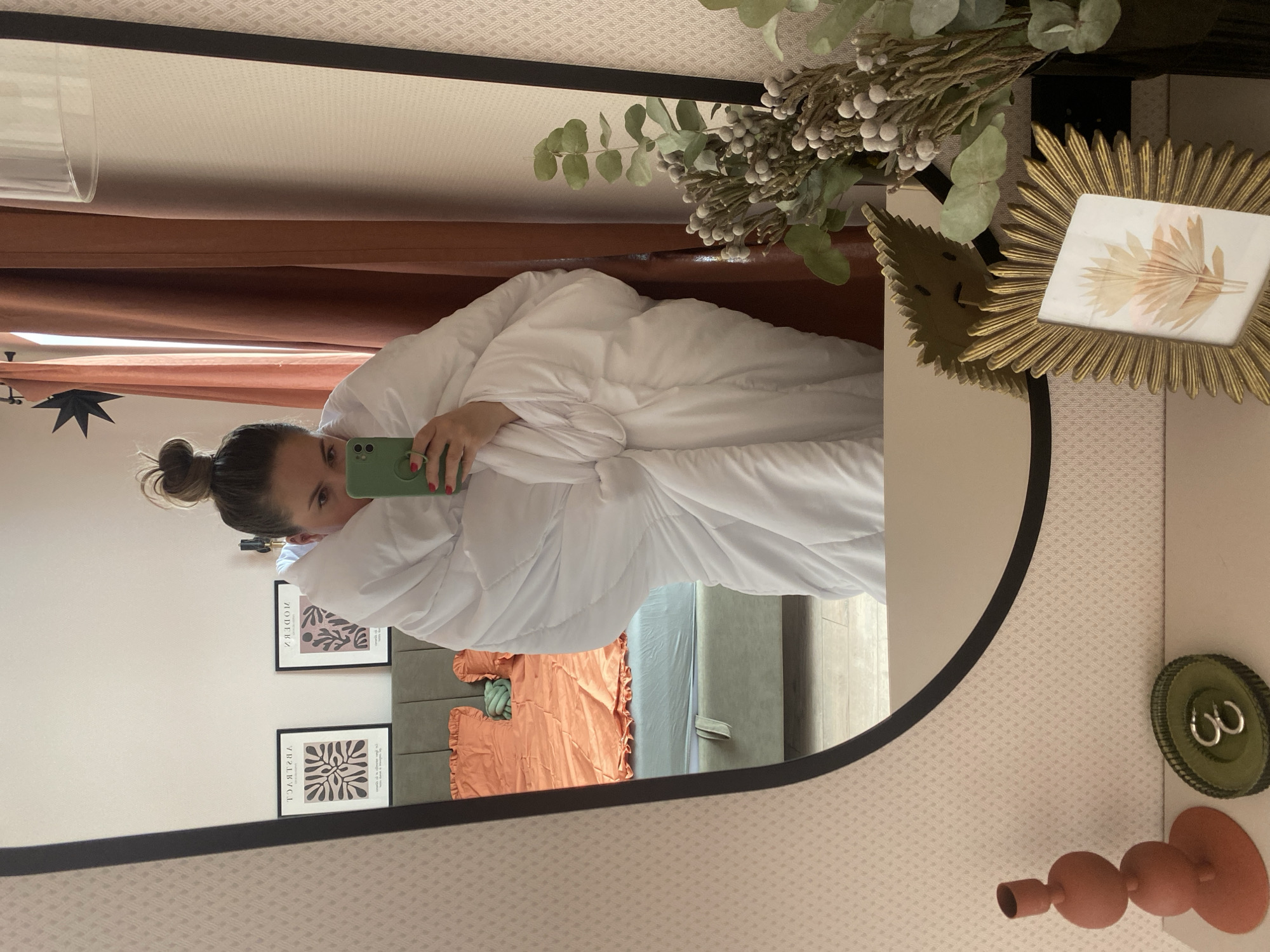 A woman is reflected in a round mirror. She is holding a green phone, partially obscured by a white duvet or comforter she is wrapped in. Her hair is in a high bun. Reflected behind her is a bed with an orange comforter and a light blue bed sheet. Two framed posters are visible on the wall behind the bed. The mirror frame is dark, and the wall around the mirror has a patterned wallpaper. A golden sunburst-shaped frame with a pressed flower inside is visible to the right of the mirror. A vase of green and white foliage is above the mirror. Other items visible on the wall to the right include green decorative rings and a terracotta-colored decorative object. There is no information available from the image to identify the location as Izhevsk, Russia, or to verify the presence of the city.