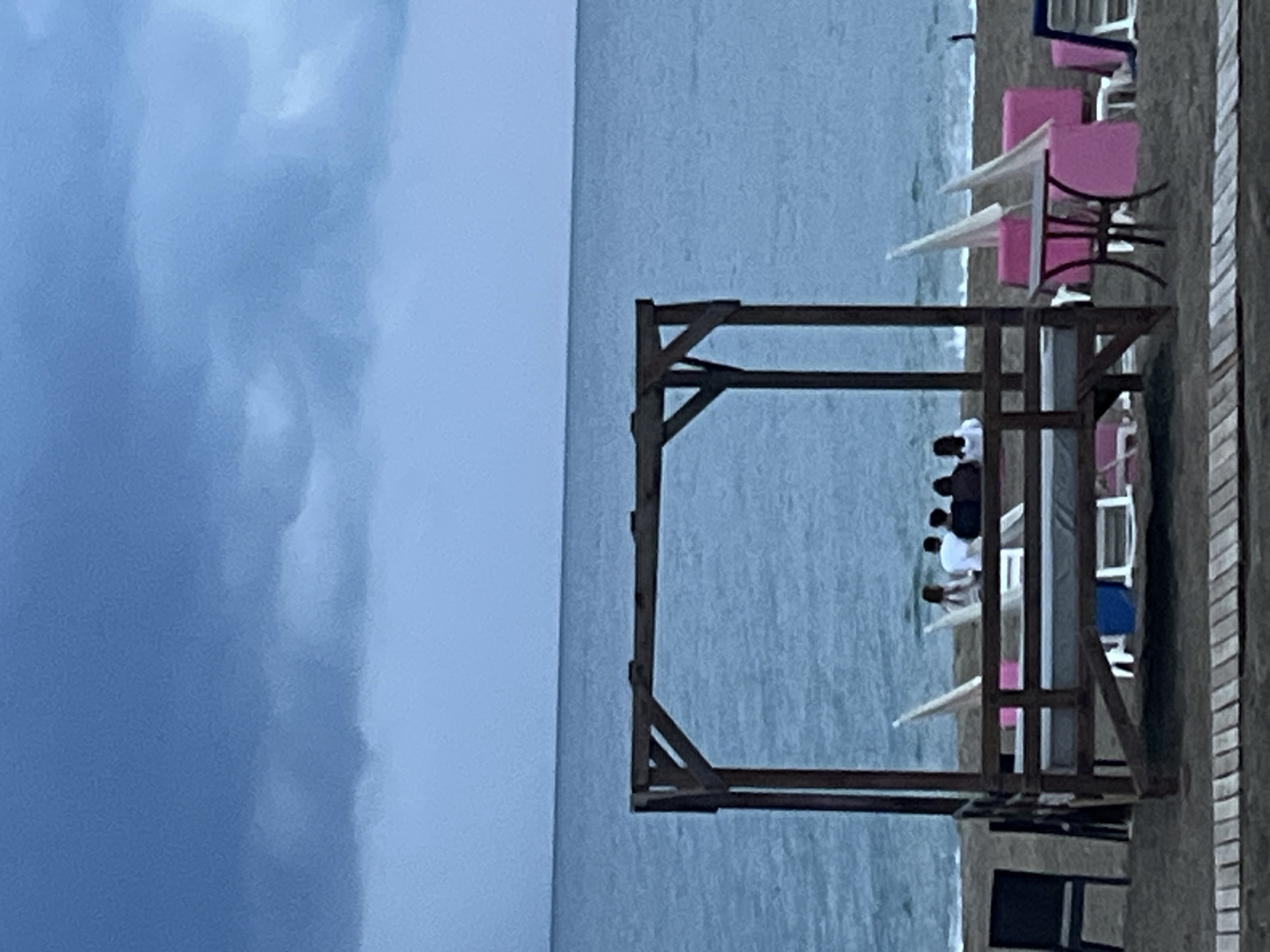The image shows a beach scene in Alanya, Turkey. A wooden structure with a canopy is erected on the sand, with people relaxing on it. Further back, pink beach chairs and tables with white umbrellas are visible. The sea occupies the middle ground of the image, and the sky is overcast with dark clouds. The photograph is taken from an elevated position, looking down at the beach.