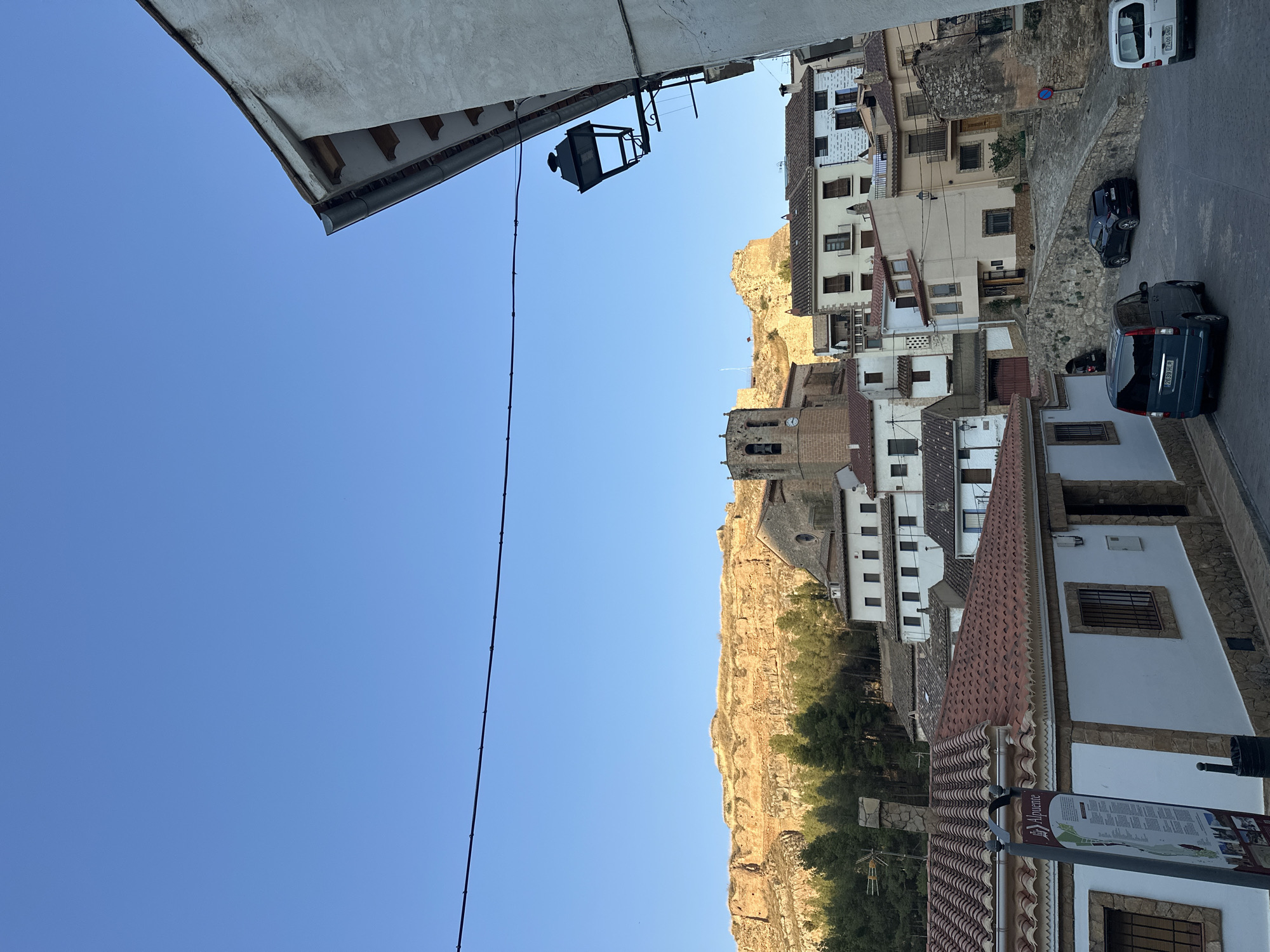 This is a horizontal, eye-level, full shot of the town of Alpuente, Spain, as seen from a street corner. The sky is a clear, bright blue. In the foreground, a white building with a stone foundation and a terracotta tiled roof runs along the bottom right of the image. A street lamp hangs from the upper right corner of the building.
To the left, a street slopes down to the right, with several parked cars visible. A dark blue minivan is parked in the lower left of the street, with license plate 2639 HLR. A black sedan is parked further up the street, and a white van is parked on the right.
The middle ground of the image features a cluster of multi-story buildings with terracotta tiled roofs. Some buildings are white, while others are painted in pastel colors like cream and light yellow. One building has a tall bell tower with a clock face.
In the background, a large, rocky cliff face rises behind the town. The cliff is bathed in golden sunlight, contrasting with the shadows cast on the town below. A line of green trees runs along the base of the cliff. A thin black wire stretches across the upper portion of the image, likely a power line or telephone wire. A signpost to the left of the foreground building has text in Spanish and an image of the town.