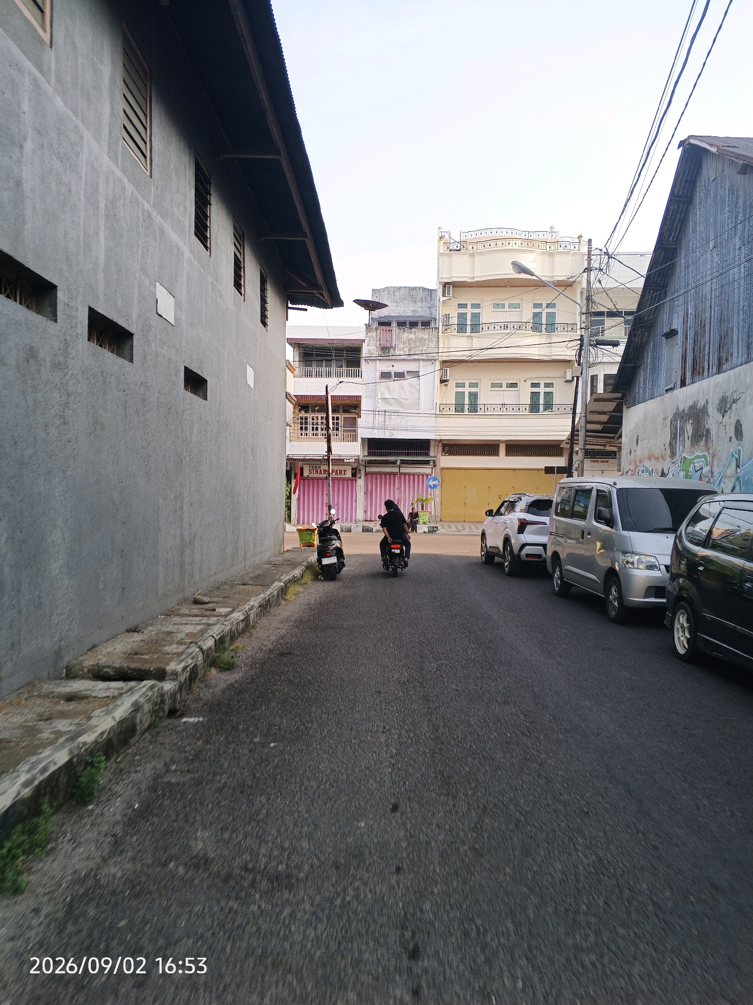 The image depicts a street scene in Pare-Pare, Indonesia, with a focus on buildings and vehicles. On the left, a gray, textured building runs along the side of the street, featuring horizontal window openings with wooden slats. The roofline is visible with corrugated metal. Across the street, a row of buildings is visible, including a multi-story structure with cream-colored walls and balconies, adorned with decorative railings. Further down the street, there are shops with pink shuttered storefronts and signage.
Several vehicles are present on the asphalt road. A black motorcycle with two riders is moving away from the camera. To the right, a silver van and a white SUV are parked. The scene is illuminated by natural light, suggesting it is daytime. Utility lines are suspended across the upper portion of the image. The date stamp at the bottom indicates "2026/09/02 16:53".