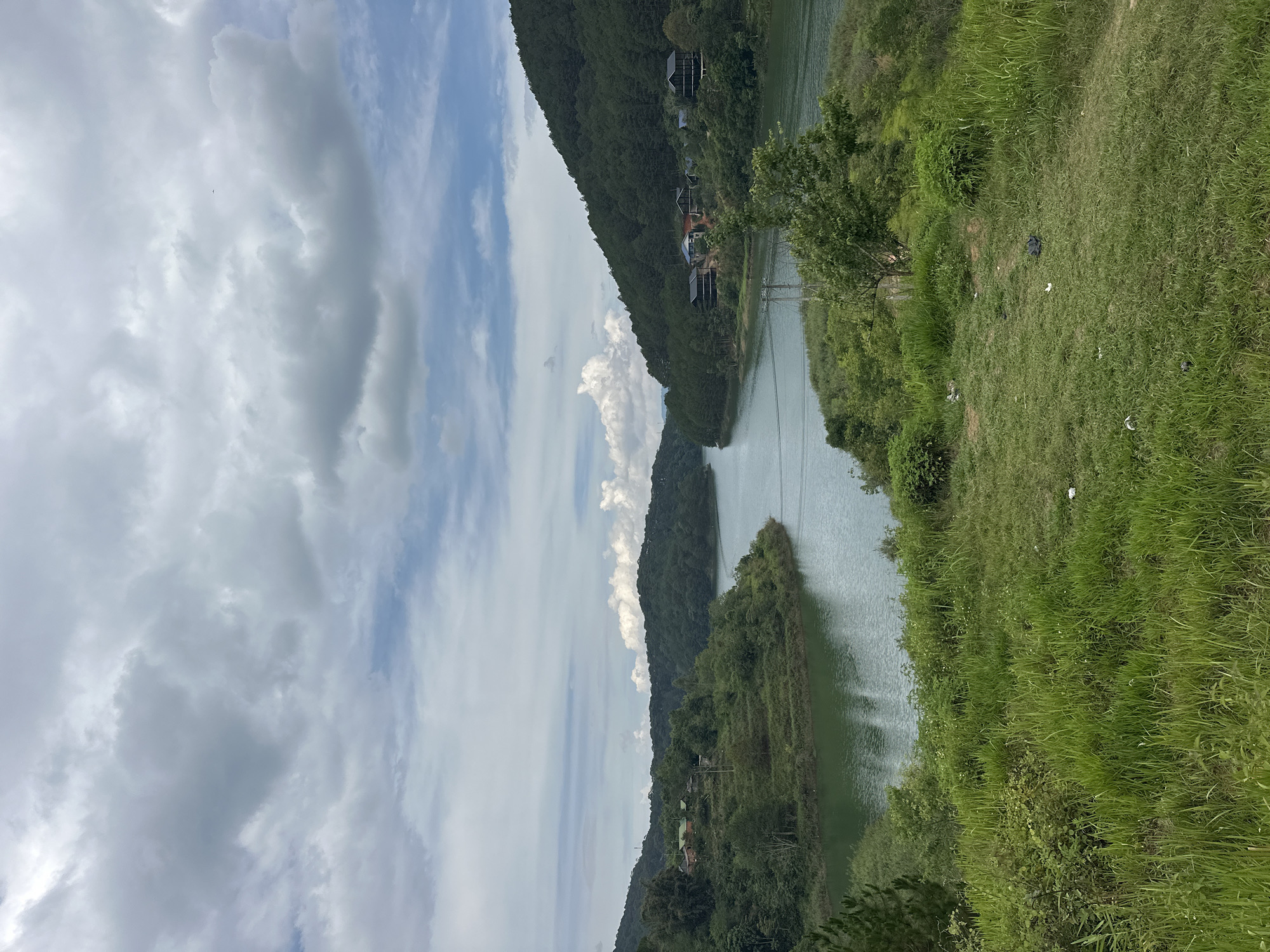 The image shows a serene view of a lake surrounded by lush green hills and vegetation. The sky is filled with clouds, ranging from puffy white formations to darker, more ominous ones. The water reflects the sky, creating a sense of depth and mirroring the cloudy expanse above. On one side, the landscape transitions to grassy areas, suggesting a closer proximity to the shore. A few structures, possibly houses or cabins, are visible along the hillside amidst the trees. The scene gives the impression of a remote, natural setting. Based on the information, the location is Da Lat, Vietnam.
