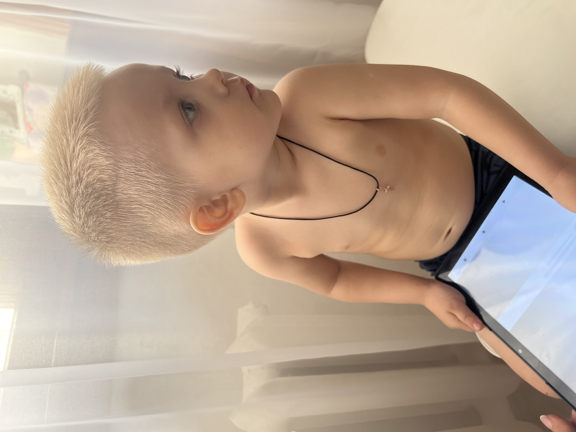 A young child, shirtless and with short, light blonde hair, is holding a tablet device. The child is wearing dark-colored bottoms and a thin necklace with a small charm. The background appears to be a softly lit room with white curtains. The location is Марковский, Russia.