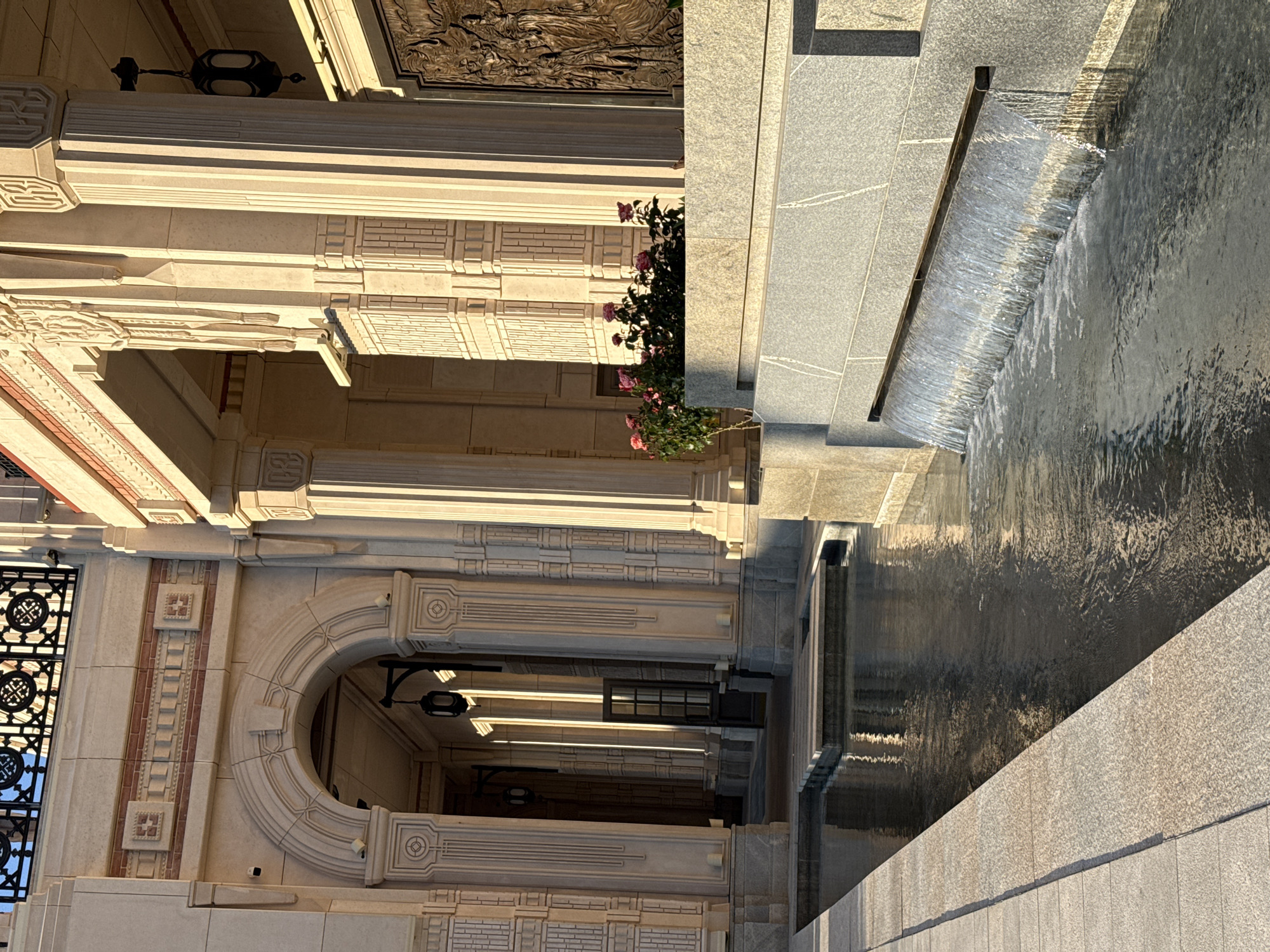 The image captures a sunny exterior view of a building with classical architectural elements, featuring large stone columns, an arched entryway, and decorative brickwork. A water feature with cascading water is prominent in the foreground, reflecting the sunlight and the surrounding architecture. A small planter with pink roses sits on a raised stone ledge to the right of the water feature. Shadows are cast across the scene, indicating the time of day. No people are visible in the image.