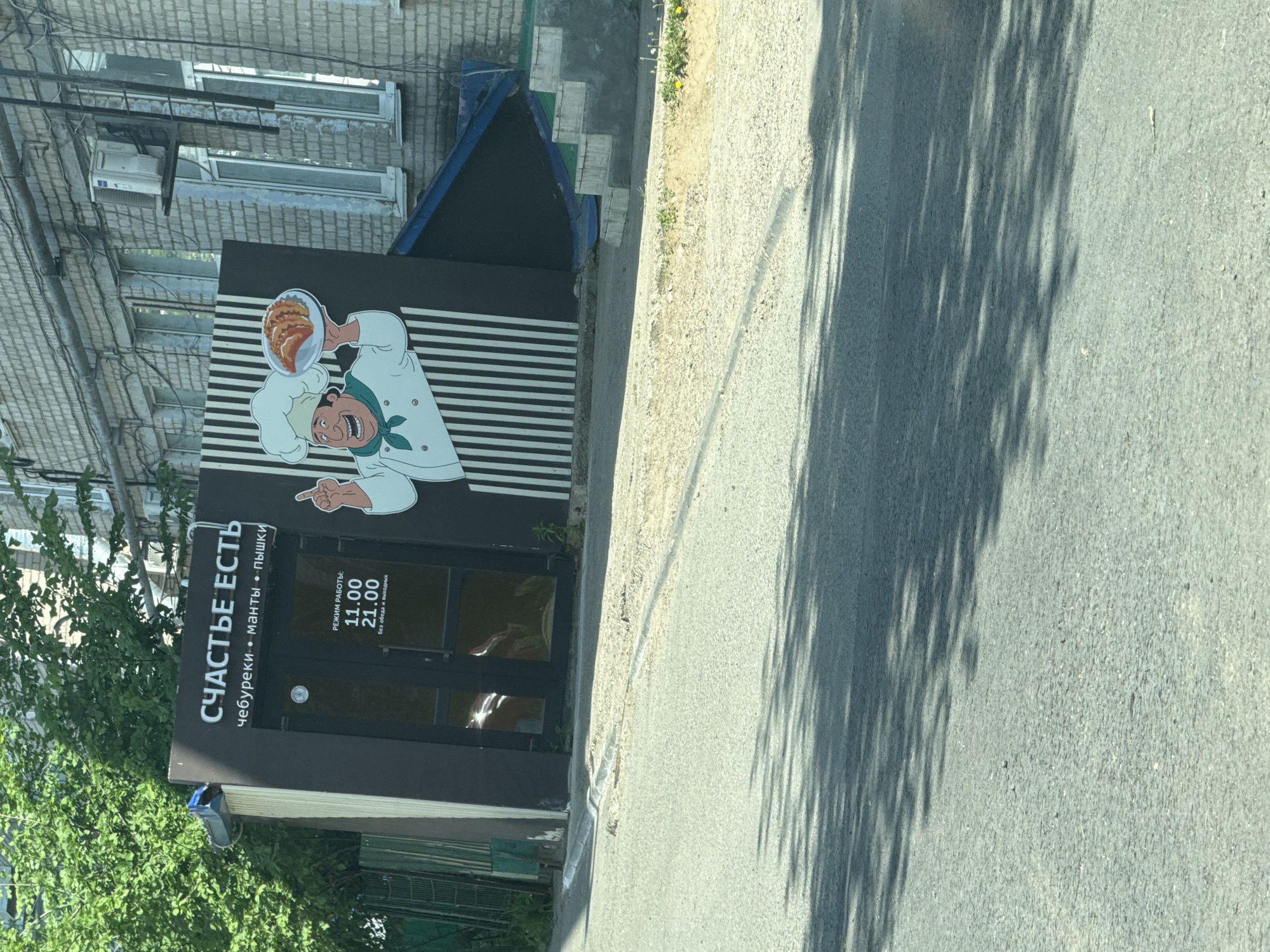 Here is a description of the image:
The image depicts the exterior of a food establishment in the city of Chaikovsky, Russia. The name of the establishment is "СЧАСТЬЕ ЕСТЬ," which roughly translates to "Happiness Exists." Below the name, it specifies the menu items: "чебуреки • манты • пышки," meaning "chebureki, manti, and pyshki."
To the left of the door is a sign indicating the operating hours: "РЕЖИМ РАБОТЫ: 11.00 - 21.00," meaning "Operating hours: 11:00 AM to 9:00 PM." The sign also notes "без обеда и выходных," indicating that it is open without lunch breaks and on weekends.
Above the entrance, there is an illustration of a smiling chef holding a plate of what appears to be chebureki. The background is striped horizontally.
The building appears to be made of brick and has windows. The surrounding environment includes foliage and a street. There are also tree shadows cast on the road.