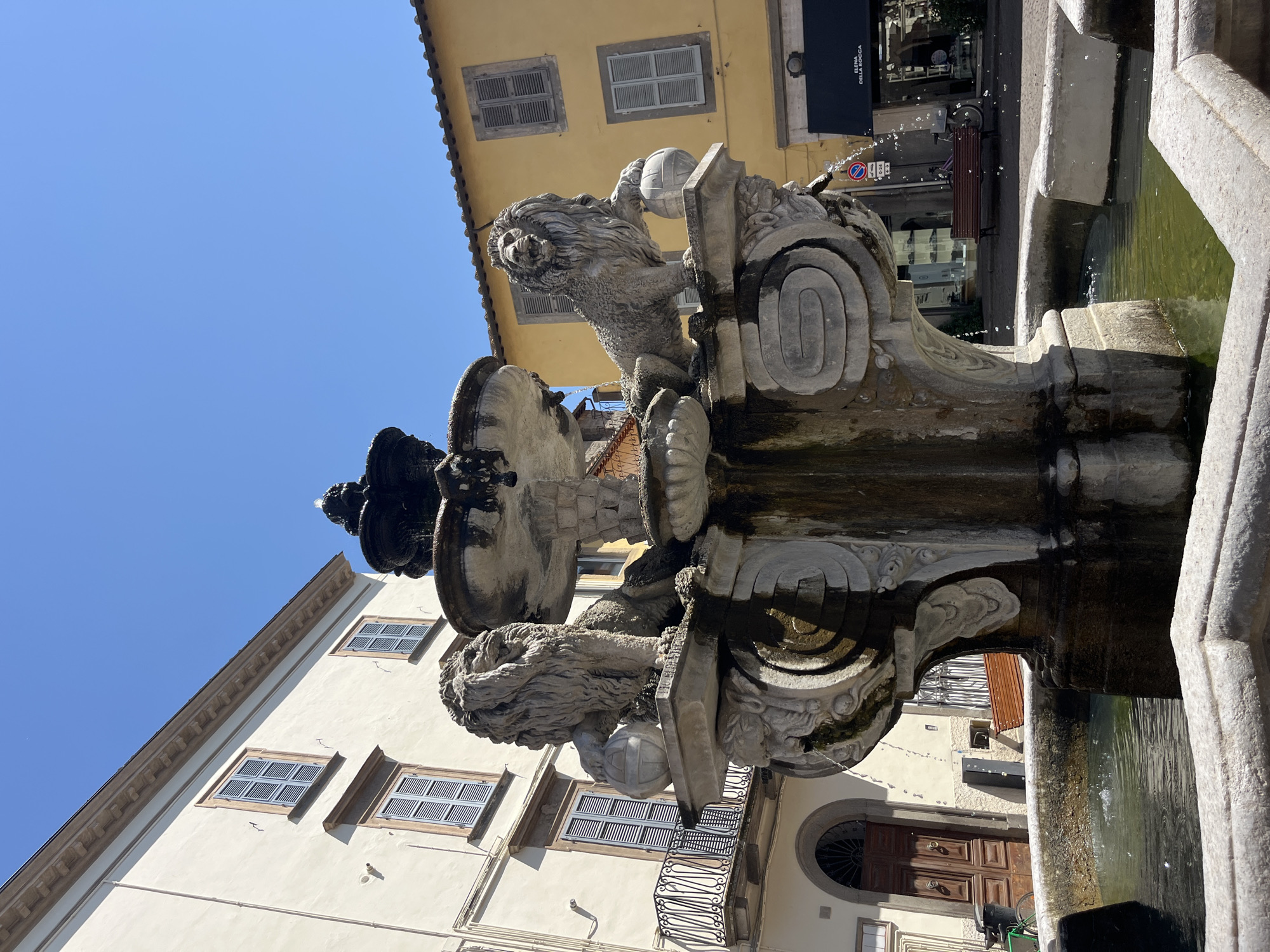 This is a full shot of a stone fountain adorned with two lion sculptures, located in the city of Viterbo, Italy, specifically in the Piazza del Plebiscito. The fountain has multiple tiers, with water gently cascading down. Each lion is positioned on a pedestal, with one paw resting on a sphere. The surrounding environment features buildings with yellow and cream facades, and blue shutters. A dark blue awning with white lettering is visible on a building to the right. A wooden bench and a bicycle are present in the background, and a small portion of a black trash bin is visible on the left. The sky is clear and blue, indicating a sunny day. There are no people actively engaged in the scene, though their presence is implied by the objects like the bench and bicycle.