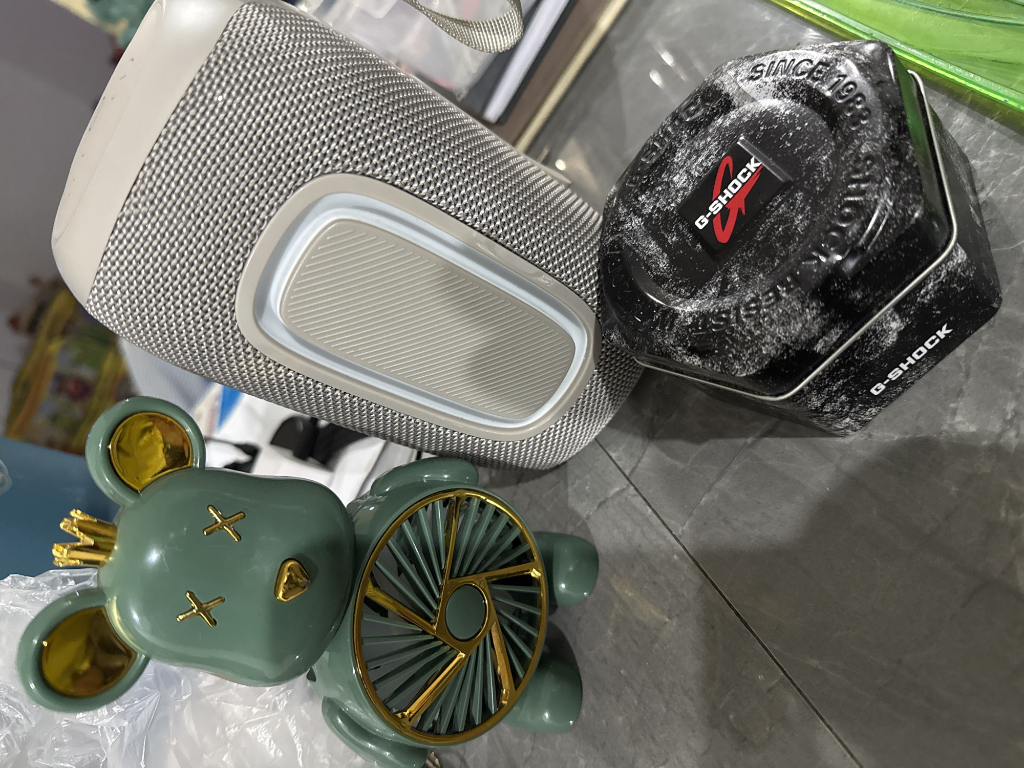 A green bear-shaped fan with gold accents and a crown sits on a gray tiled surface next to a beige and gray cylindrical speaker. In the foreground, a black G-Shock watch box with distressed white markings and the G-Shock logo is visible. The lighting suggests an indoor setting. The image does not provide any information about the location of Apostadero, Venezuela.