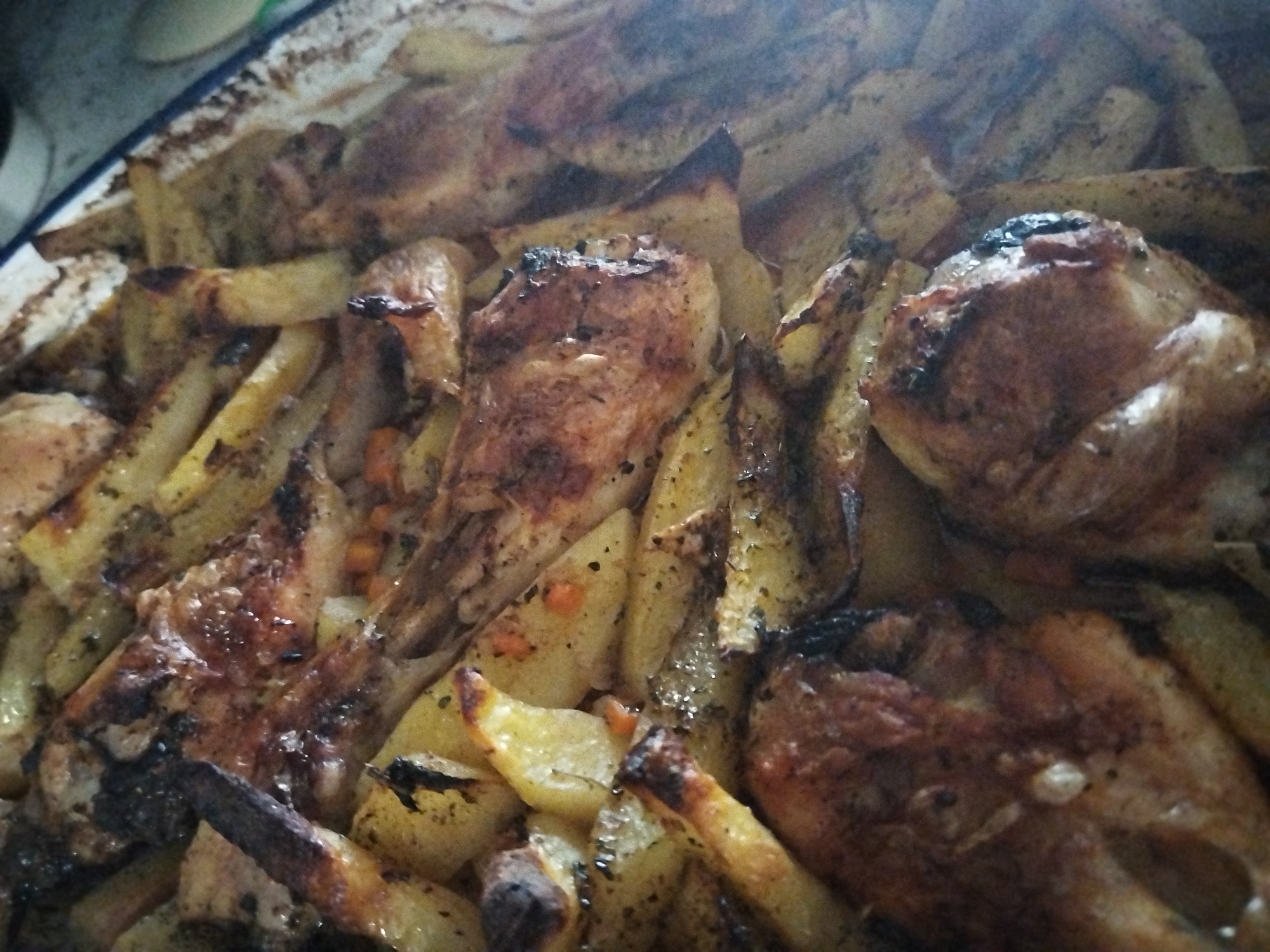 The image depicts a baking tray filled with roasted chicken and potatoes. The chicken pieces are browned and appear well-cooked, with some parts showing darker, potentially burnt areas. The potatoes are cut into irregular shapes and are also roasted, exhibiting a golden-brown color. The dish seems to be seasoned with herbs and spices. The environment is indoors, with the baking tray possibly on a kitchen counter or table.