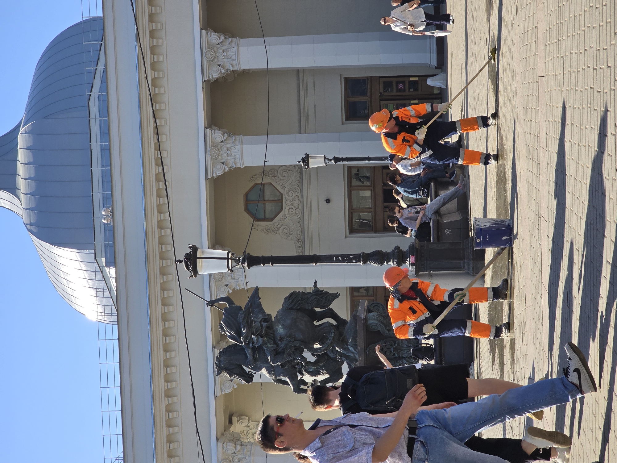 The image captures a sunny day in Moscow, Russia, near a building with a distinctive dome. Two workers in orange safety gear clean the pavement with long-handled tools, while passersby, some seated, observe. A bronze sculpture of horses and riders stands near a lamppost. The architecture of the building features columns and decorative elements.