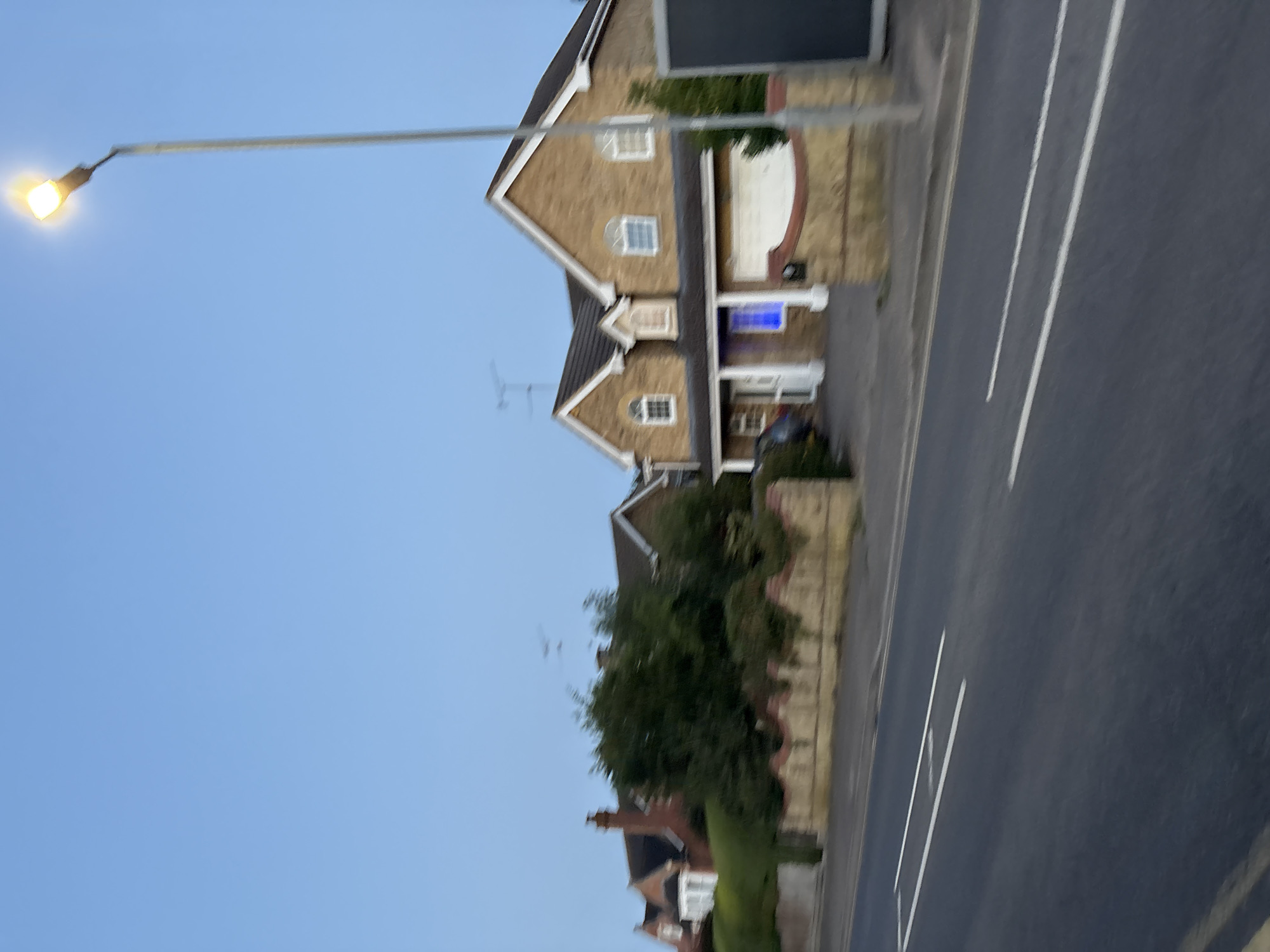 This image captures a suburban street scene in Chelmsford, United Kingdom, under a clear twilight sky. A road with white line markings is prominently featured in the foreground, suggesting it is a typical residential street. To the right of the road, a tall street lamp illuminates the darkening sky. Beyond the road and separated by a low, decorative brick wall topped with greenery, are residential properties. The houses appear to be detached or semi-detached, constructed with brick and featuring pitched roofs. One house, in particular, is illuminated with a bright blue light emanating from an interior window. The overall impression is of a quiet, residential area as dusk settles.