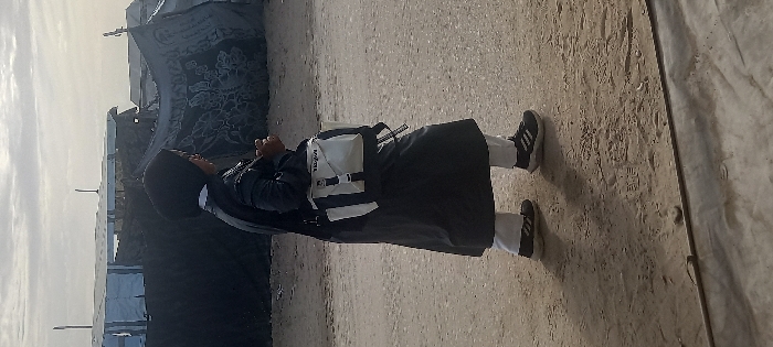 The image depicts an individual walking from left to right across an uneven dirt path in Khan Yunis, Palestinian Territory. The person wears a black hijab, a dark, long-sleeved outer garment, white pants, and black athletic shoes with white accents. A white and black shoulder bag, labeled "TROJAN", is carried across their body. In the background, a cluster of rudimentary shelters or tents is visible, constructed from tarpaulins, some blue and white, others dark and patterned, set against a cloudy, overcast sky. The ground is consistently light brown dirt with small debris.