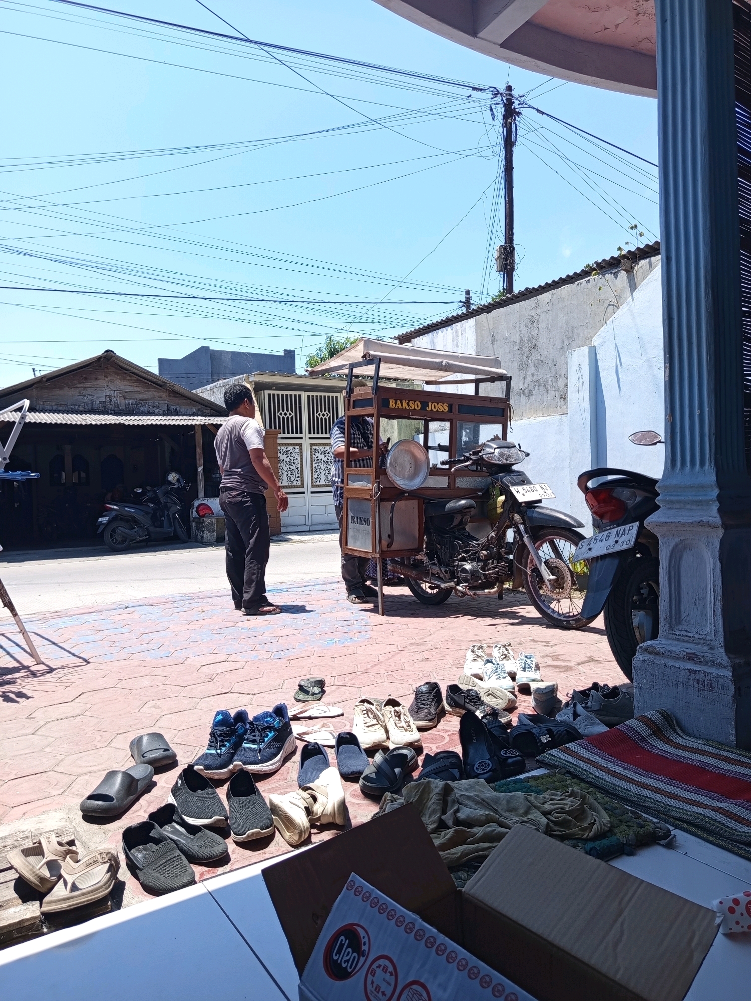 This is a daylight, medium shot photograph taken from a slightly elevated angle. The scene depicts a street in Ngimbangan, Indonesia. In the foreground, a variety of footwear, including sandals, flip-flops, and sneakers, are arranged on a patterned paved surface. Further back, a man in a dark t-shirt and black pants stands near a food cart with the text "BAKSO JOSS" visible. A motorcycle is parked next to the food cart. In the background, there are buildings with corrugated metal roofs, a motorcycle parked under a shed-like structure, and a utility pole with numerous wires. The sky is clear and blue.