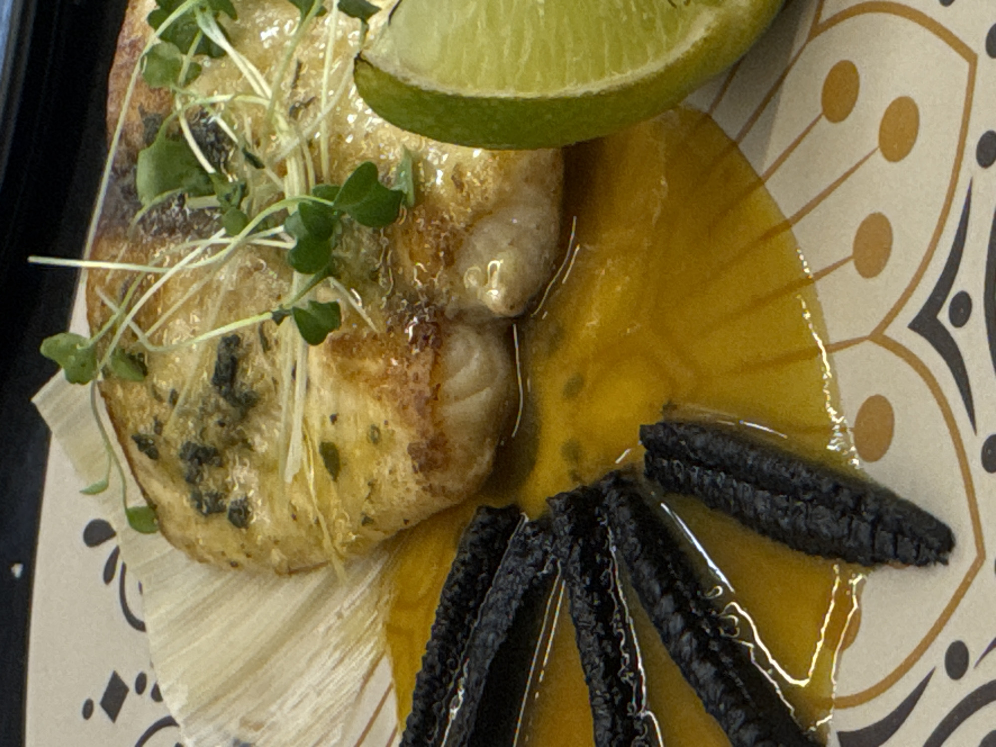 This close-up shot features a plated dish of food. The main subject is a piece of light-colored fish, possibly cod or haddock, that has been pan-seared or baked to a golden-brown hue. It is topped with fresh green microgreens, adding a delicate, earthy contrast. The fish rests on a decorative plate with a cream background and an intricate, abstract floral pattern in shades of brown and black.
Surrounding the fish is a vibrant, translucent yellow sauce, likely a citrus-based coulis or a fruit puree, with a glossy sheen. A slice of lime, with its bright green rind and white pith, is perched on top of the dish, suggesting a zesty flavor profile. In the foreground, fanned out in a semi-circle, are several dark, elongated pieces of food. These appear to be charred or grilled corn kernels or perhaps a type of black bean crisp, adding a textural and visual element.
The setting is a restaurant or a home dining environment, given the presentation of the food on a formal plate. The lighting is bright and even, highlighting the textures and colors of the dish. There is no visible text in the image, and the time of day and weather are not discernible. The focus is entirely on the culinary composition. The dish evokes a sense of a carefully prepared meal, likely from a modern cuisine establishment in Sofia, Bulgaria.