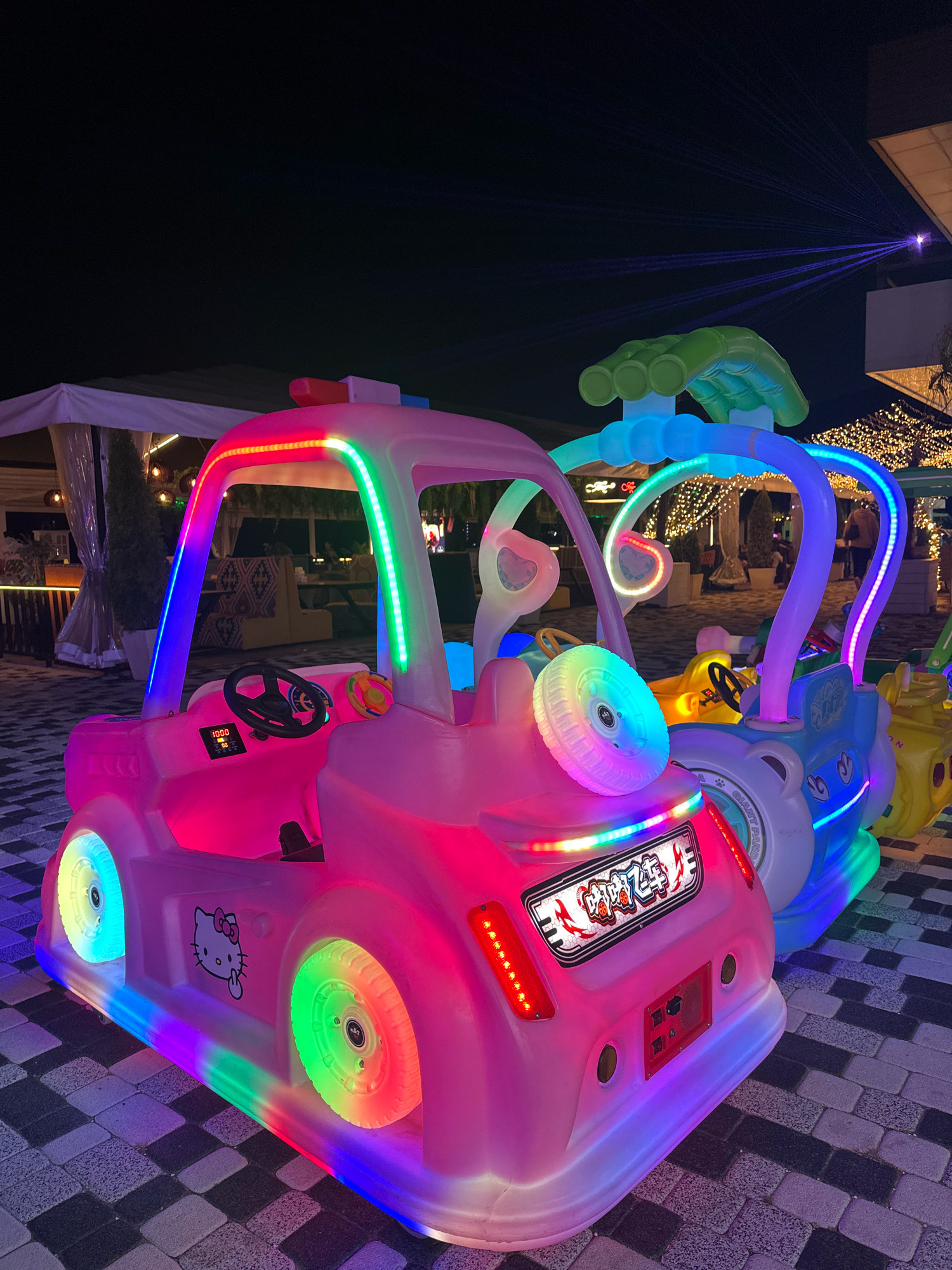 A nighttime outdoor scene features several brightly lit children's amusement rides designed to resemble cars. The ride in the foreground is predominantly pink and is adorned with colorful LED lights along its frame, wheels, and accents. It has a Hello Kitty decal on its side and a digital display on its dashboard showing "1000" and "20". Behind it, a blue and white ride is partially visible, also illuminated with LEDs, and further back, yellow and other colored rides can be seen. The rides are situated on a paved surface with a checkered pattern of dark and light gray stones. In the background, there are tents with white canopies, outdoor seating areas with tables and chairs, and numerous string lights creating a festive atmosphere. A faint blue laser light display is visible in the upper right portion of the image. The location is identified as Lermontovo, Russia.