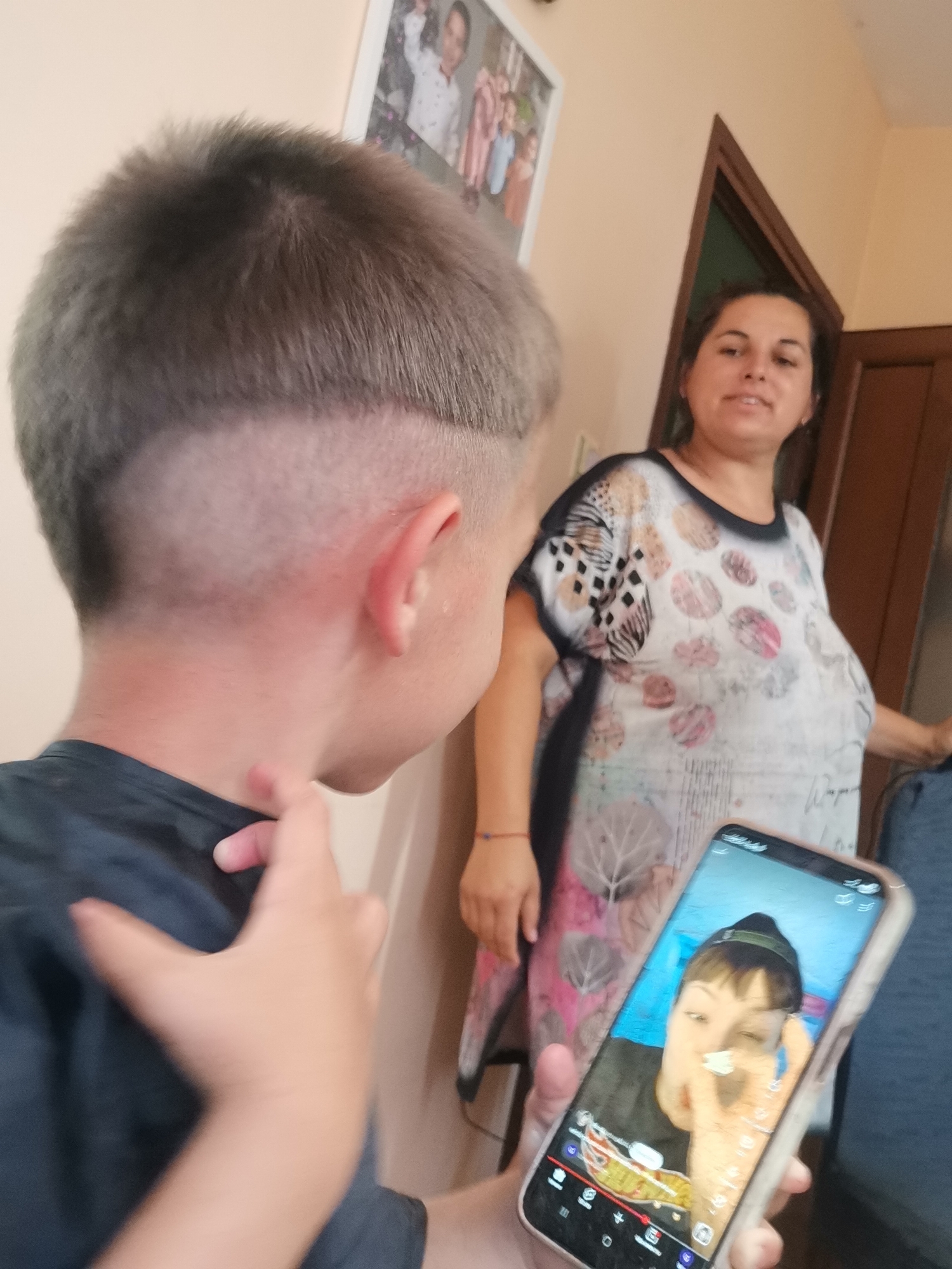 This is a vertical, indoor, eye-level shot of a young boy in the foreground, his back and the side of his head facing the camera. He has a freshly cut haircut with a buzzed lower back and sides, and longer hair on top. His left hand is visible, with his index finger extended and touching his neck. He is wearing a dark t-shirt.
In the mid-ground, a woman stands to the right of the boy. She is wearing a white t-shirt with a colorful, abstract pattern of circles and leaves. She is looking towards the boy and appears to be smiling.
In the foreground, a smartphone is held in the boy's right hand, angled towards himself. The screen displays a selfie video or photo of a person wearing a green beanie and a black t-shirt, making a peace sign with their fingers. The user interface of a social media app, possibly TikTok, is visible on the phone screen with icons and text.
The setting appears to be a home, with a plain wall behind the subjects and a partially visible doorway with dark wood trim. A framed picture of a family is visible on the wall above the woman. The lighting suggests it is daytime. The overall emotion conveyed is casual and familial.