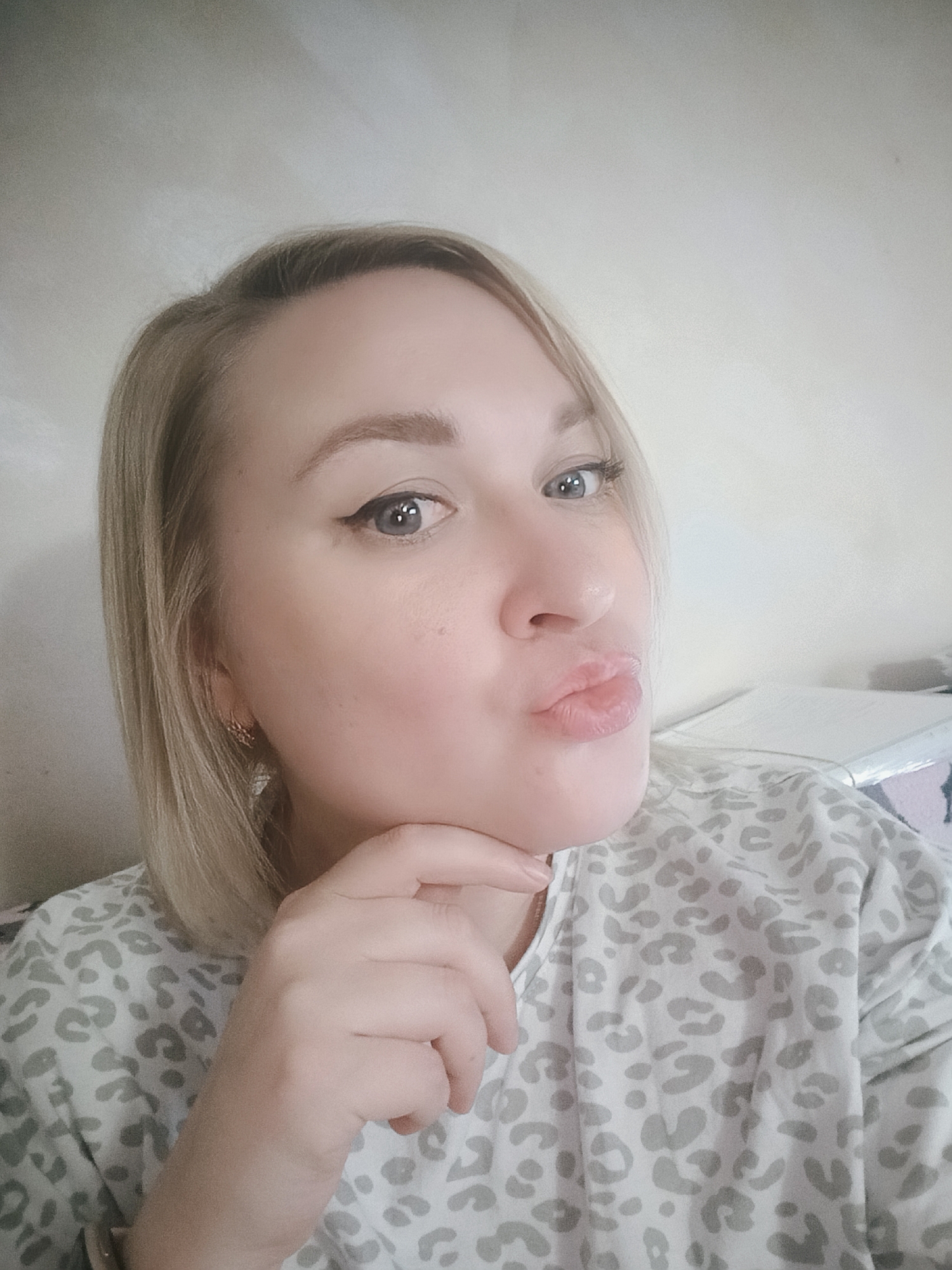 A blonde woman with fair skin is posing for a selfie. She is wearing a white, short-sleeved shirt with a gray leopard print. Her hair is short and styled, and she has on eyeliner. She is making a kissing face and has her hand under her chin. The background is a plain, light-colored wall. The location is unknown.