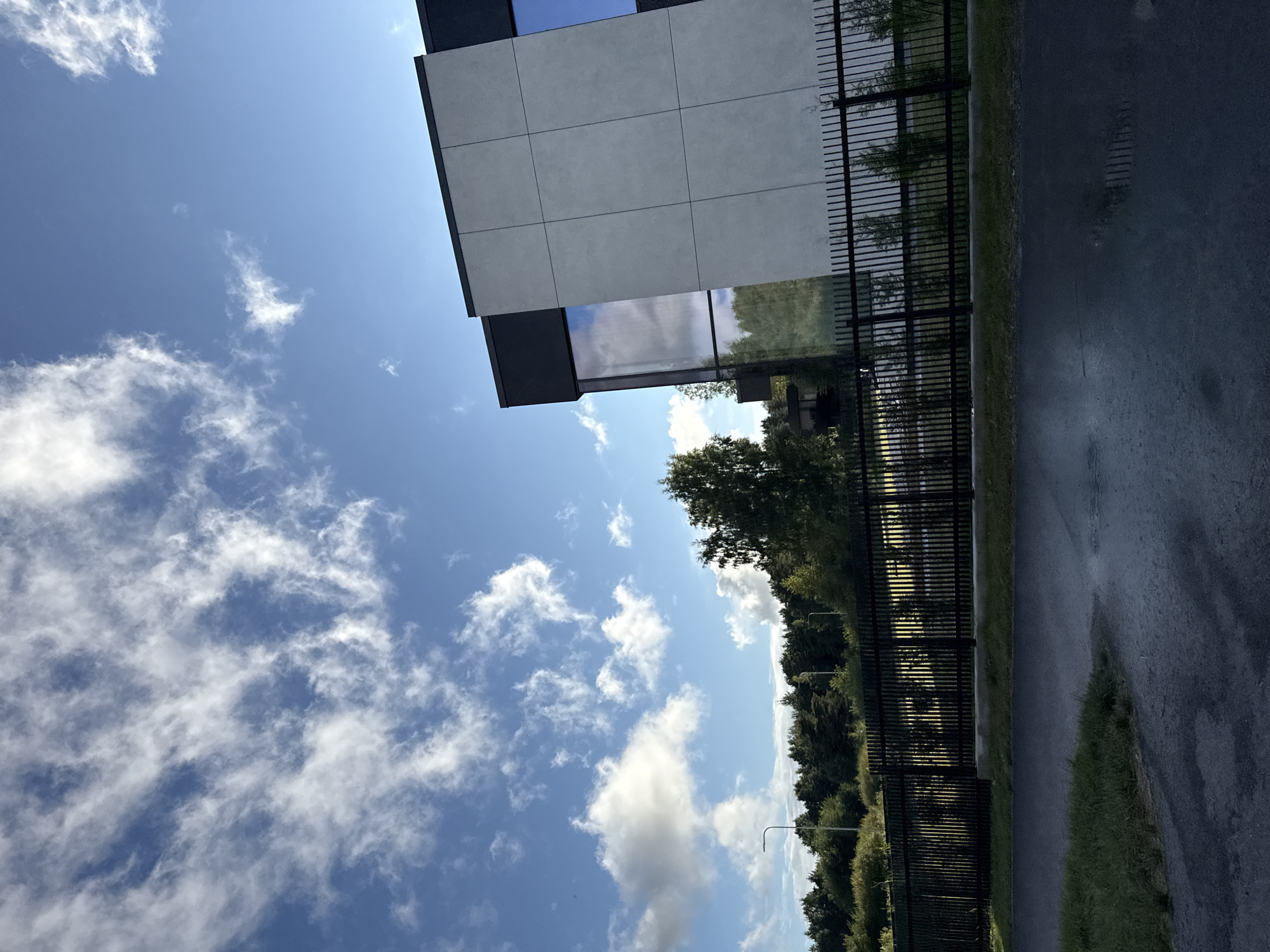 In Harkujärve, Estonia, a modern building with a grey tiled facade and dark accents stands against a bright blue sky dotted with scattered white clouds. A black metal fence runs along the foreground, separating a grassy area from a road. Beyond the fence, a dense line of trees forms a backdrop, with a few taller trees visible on the right side of the image. The road in the foreground appears wet, suggesting recent rainfall. The scene depicts an exterior view of a commercial or residential property in a semi-rural setting.