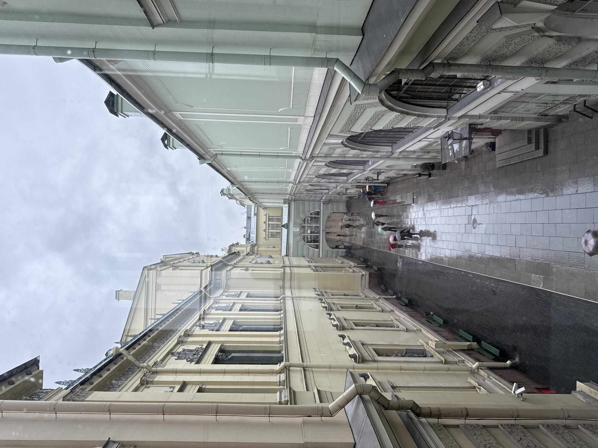 The image depicts a rainy day in a courtyard between two buildings in Saint Petersburg, Russia. The buildings, characterized by their classical architectural style with ornate facades, are painted in pale yellow and pale green. The courtyard is paved with stone tiles, which are wet and reflective due to the rain. Several people are visible walking with umbrellas, indicating the inclement weather. One person is seen holding a dark umbrella, while others are using colorful umbrellas in shades of red, pink, and green. The scene appears to be an outdoor passage or a courtyard within a historical district or a museum complex, given the grandeur of the architecture. The sky is overcast, contributing to the somber atmosphere of the rainy day. In the foreground, a security camera is mounted on the pale green building, and green benches are visible along the pale yellow building. The overall impression is one of a damp, yet architecturally rich urban environment.