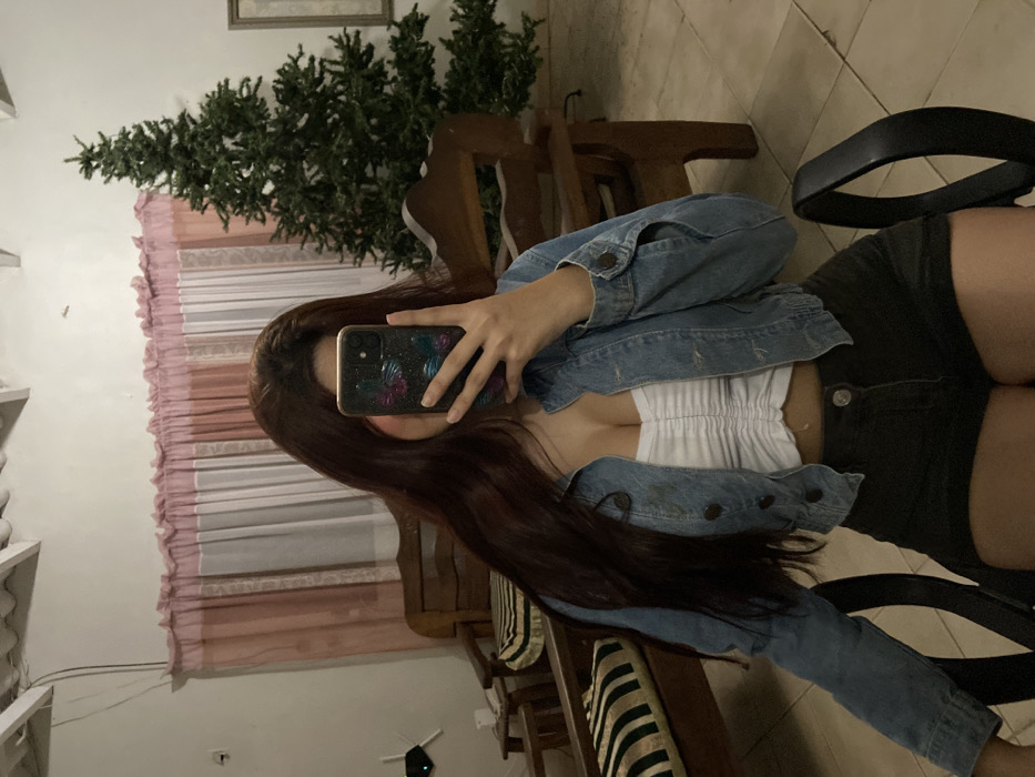 A woman takes a selfie in a mirror, holding her phone in front of her face. She is wearing a light blue denim jacket over a white top and dark shorts. Long brown hair cascades down. The reflection shows a room setting with a Christmas tree, wooden chairs with striped cushions, and pink-toned curtains. A black office chair is also visible in the scene.