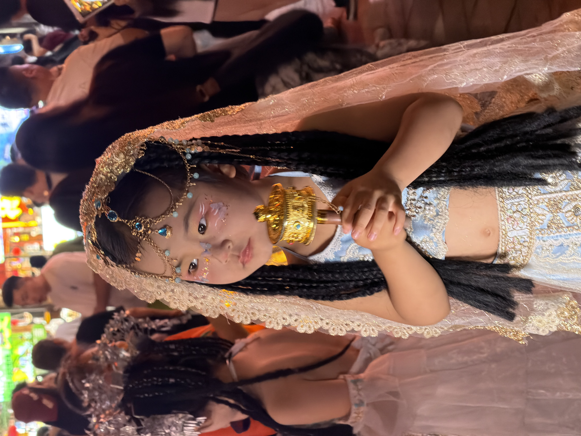 In Yuzhong District, China, at night, a young girl is dressed in ornate cultural attire, featuring a golden headpiece with delicate chains and blue gemstones, a light blue cropped top with intricate gold embroidery, and a matching skirt. Her dark hair is styled in long braids that frame her face, which is adorned with light makeup and a subtle butterfly design. She holds a golden prayer wheel in her hands. To her left, another child is visible, also in elaborate white and pink layered clothing, with a silver headdress. The background is blurred, showing a crowded street scene with people in casual clothing and various colorful signs, suggesting a lively night market or festival environment.