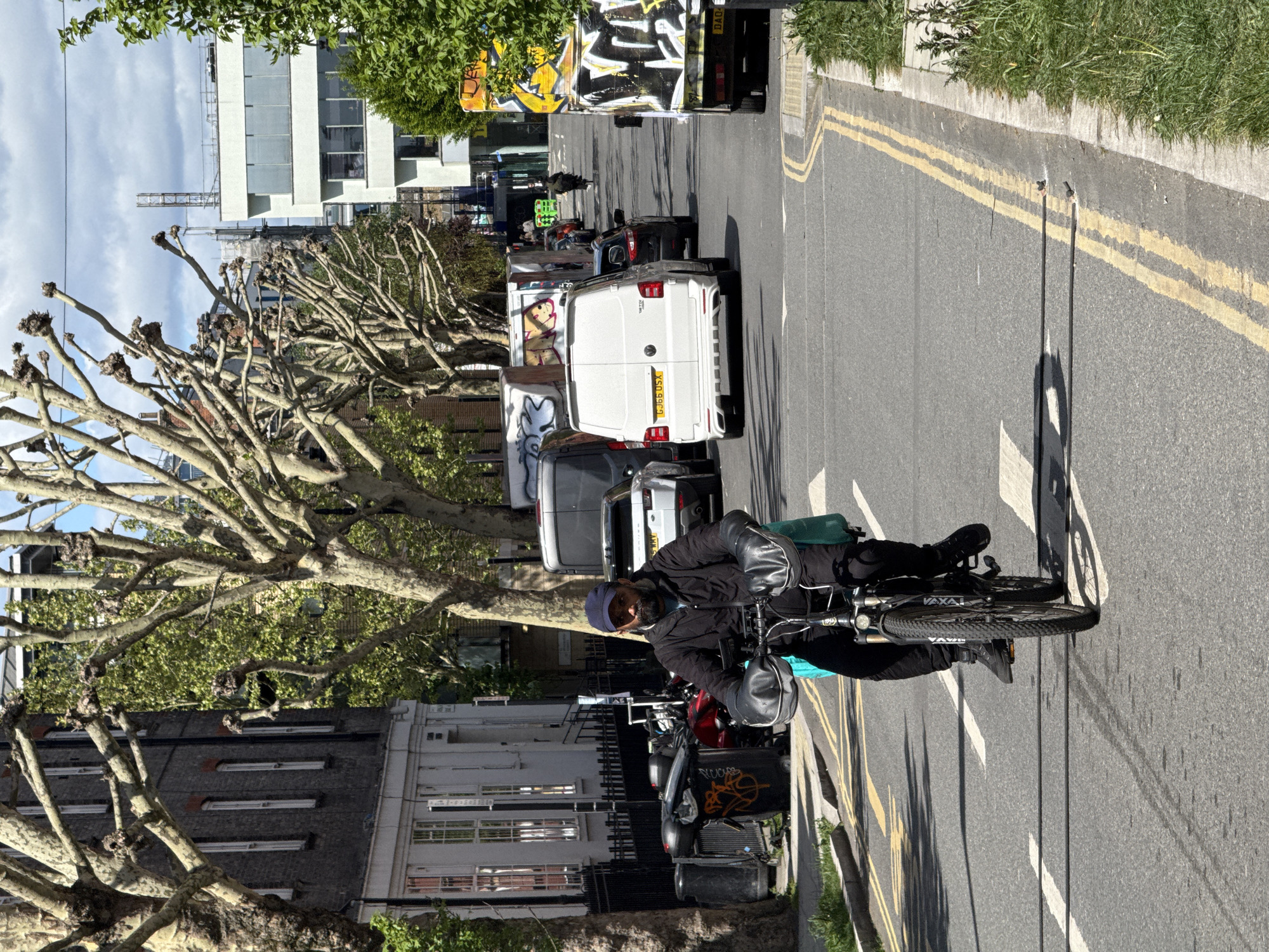 The image depicts a street in London, United Kingdom, with a cyclist riding on the right side of the road. The road is marked with double yellow lines and white arrows. There are several vehicles parked on the left side of the road, including vans and a truck with graffiti. Buildings line the left side of the street, with trees planted along the sidewalk. The cyclist is wearing a dark jacket, gloves, and a blue cap. The sky is overcast. A construction crane is visible in the distance.