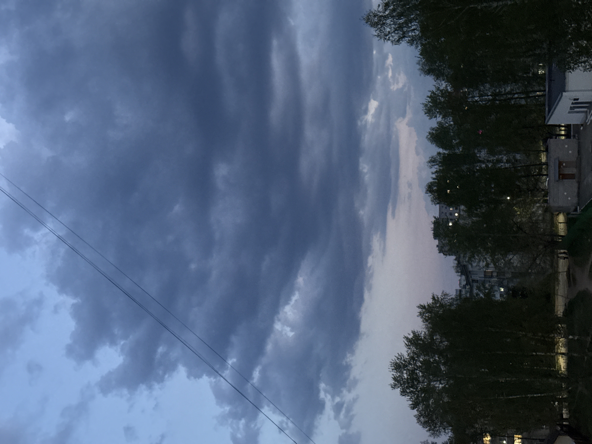 The image depicts a view of the sky with dark, ominous clouds dominating the upper left portion of the frame. A set of electrical or utility wires stretches diagonally across the bottom left. Along the right side of the frame, there's a row of trees with dense foliage, obscuring parts of buildings. Some windows are visible in the buildings, suggesting residential or commercial structures. The overall lighting suggests it is dusk or dawn. The location seems to be an urban area with buildings and greenery, but the specific location in the city of Unknown, Unknown is unidentifiable based on the visual information alone. No people are directly visible in the frame.