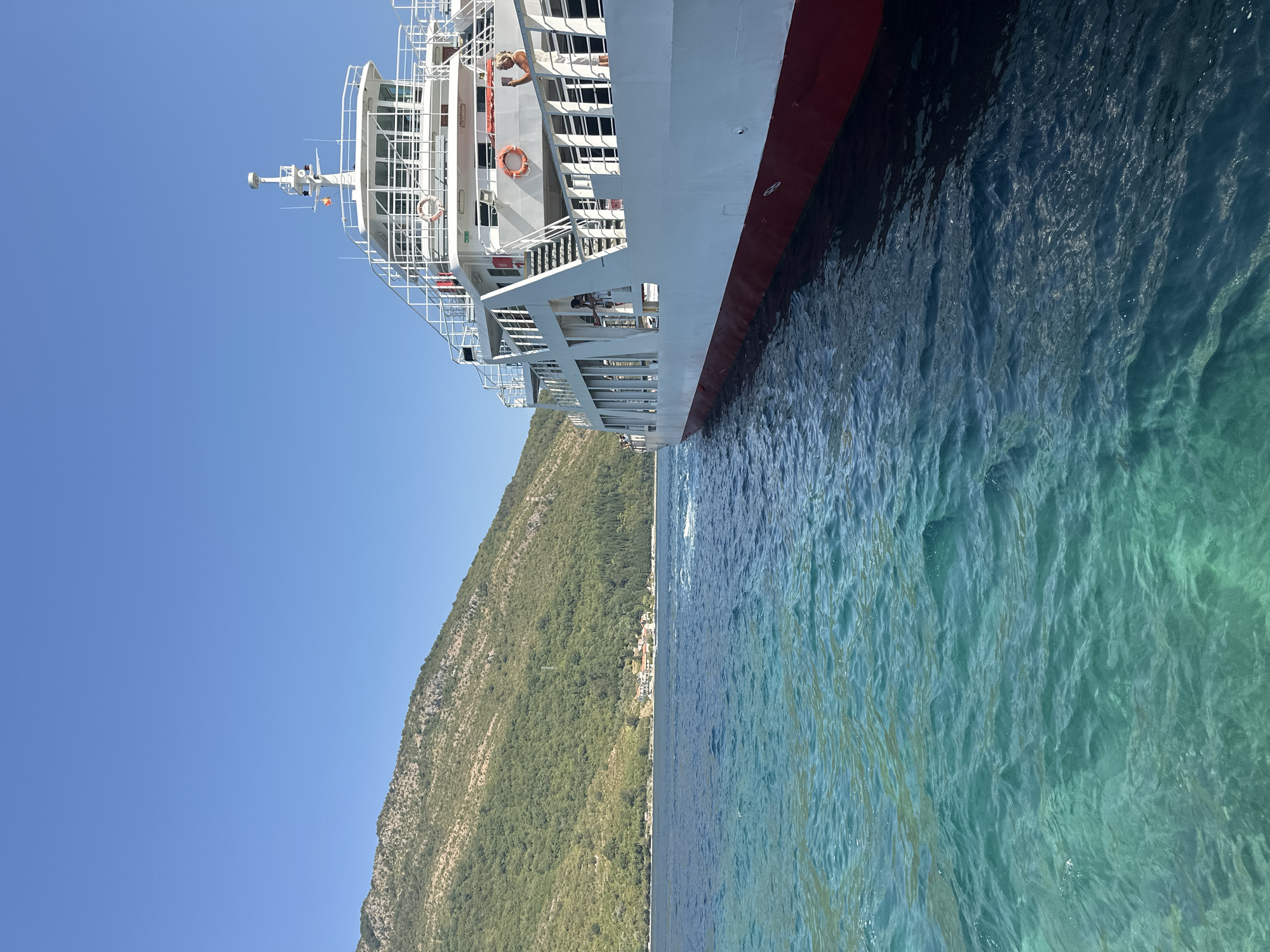 An exterior, eye-level, medium shot captures a ferry sailing on the clear, turquoise waters of the Bay of Kotor, near Lepetane, Montenegro. The ferry, predominantly white with a red waterline, occupies the right side of the frame, its upper decks and navigation bridge visible. The left side of the image showcases a lush, green, tree-covered mountain rising steeply from the water's edge, with a small cluster of buildings nestled on its lower slopes. The sky above is a clear, bright blue. Several passengers are visible on the ferry's decks, including one woman taking a photo with her phone. The water is choppy, with small waves creating ripples and white foam trails behind the vessel. The scene conveys a sense of serene travel under a sunny sky.