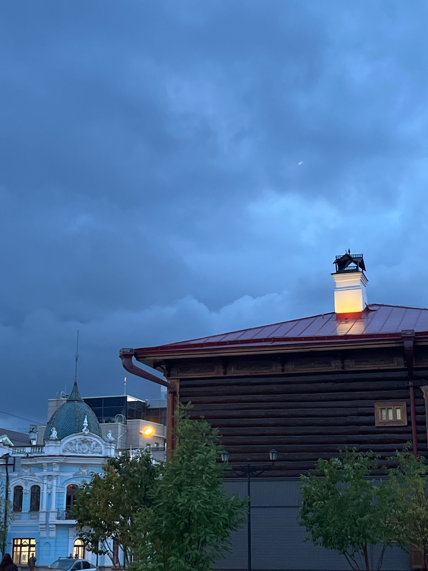 The image depicts a twilight scene in the city of Krasnoyarsk, Russia, likely on Teatralnaya Square. In the foreground, a traditional wooden log building with a red metal roof is prominent. A white chimney with decorative dark metalwork on top stands on the roof, illuminated from within by a warm light. To the left of the wooden building, a light blue, ornate building with a green dome and intricate white stucco details is visible. The sky is cloudy and dark blue, with a faint crescent moon visible. Several trees with green foliage are scattered throughout the scene. In the background, modern buildings with illuminated windows can be seen, including a taller glass-fronted structure. The street level shows parked cars and a lamppost. The overall atmosphere is moody and calm, characteristic of dusk in a city.